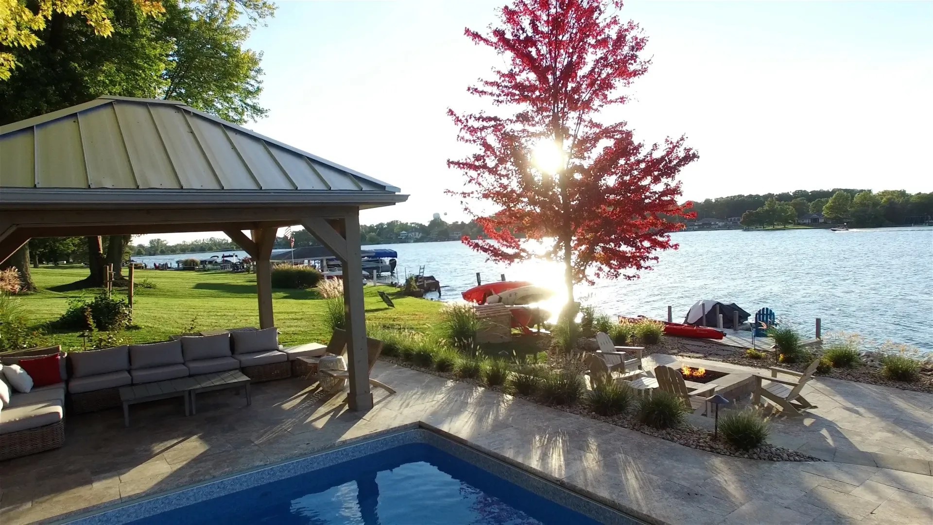 Gazebo with seating overlooking a lake, a pool in the foreground, and a tree with red leaves illuminated by the sun.