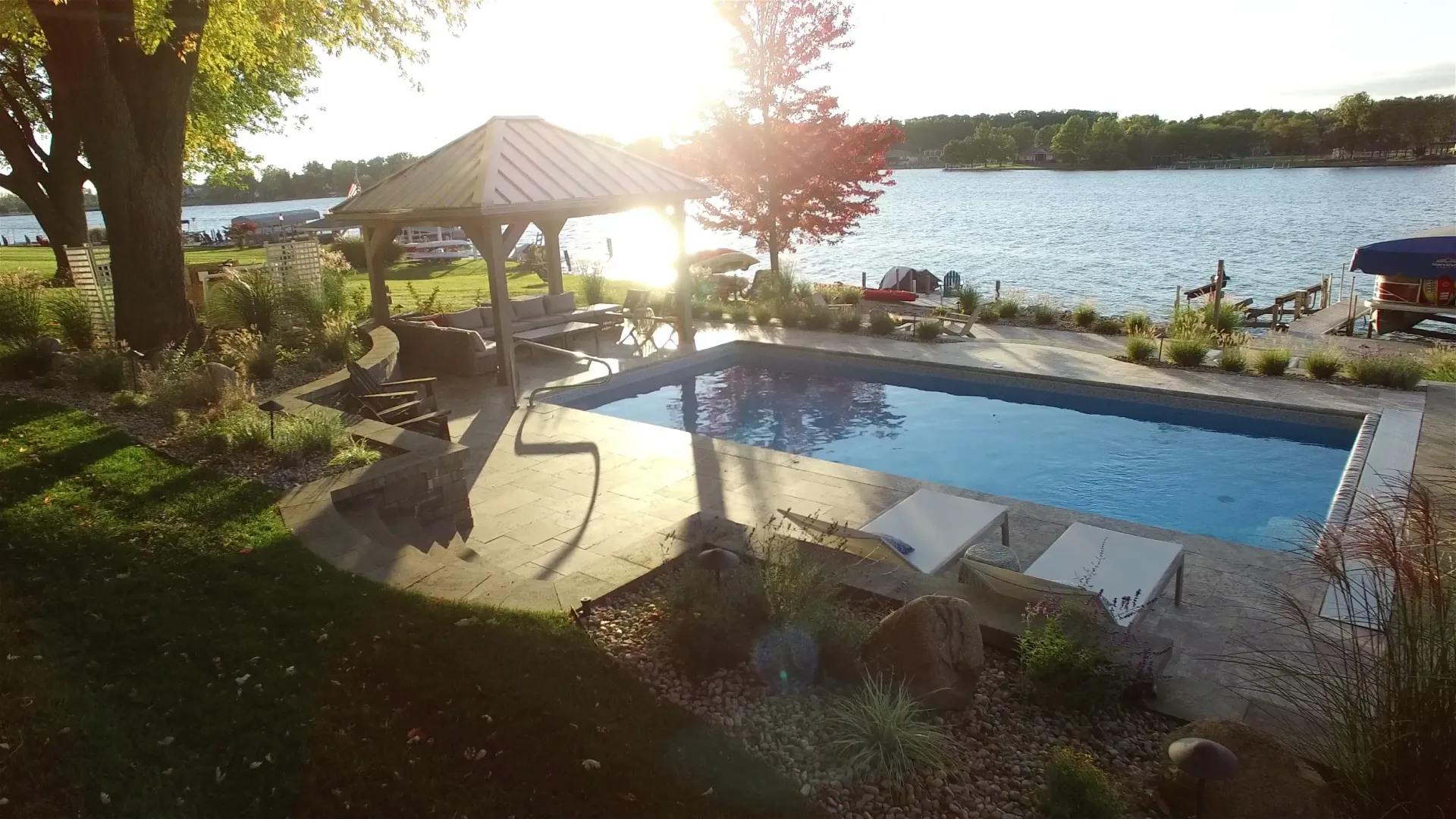 A rectangular pool next to a lake, with a shaded seating area and a setting sun.