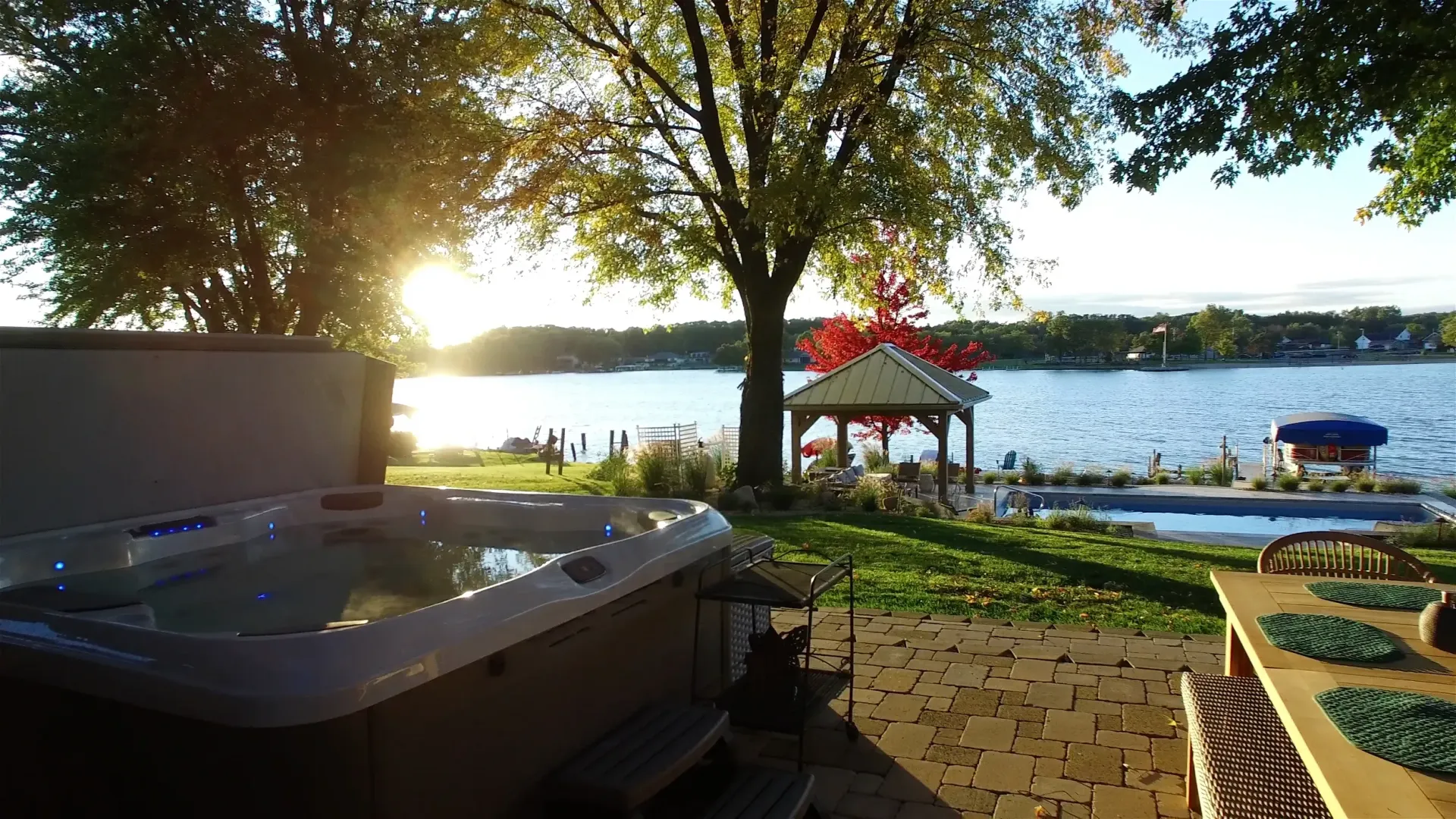 A hot tub overlooks a lake at sunset. A gazebo and table are visible on the grassy lawn.