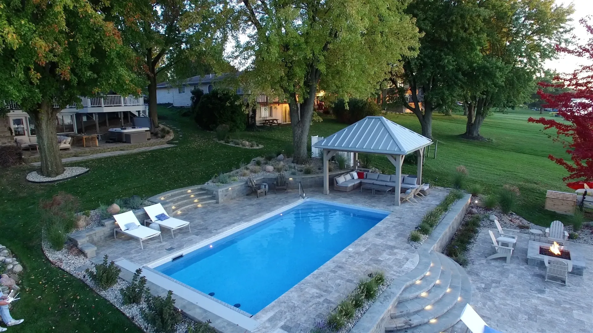 An aerial view of a backyard with a rectangular pool, gazebo, and seating areas surrounded by trees and green grass.