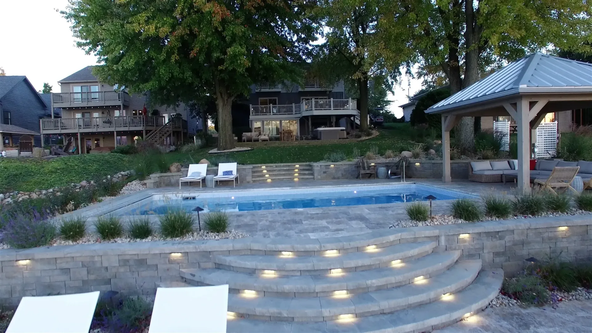 A stone patio with a pool and gazebo near waterfront houses; steps with lights lead down.