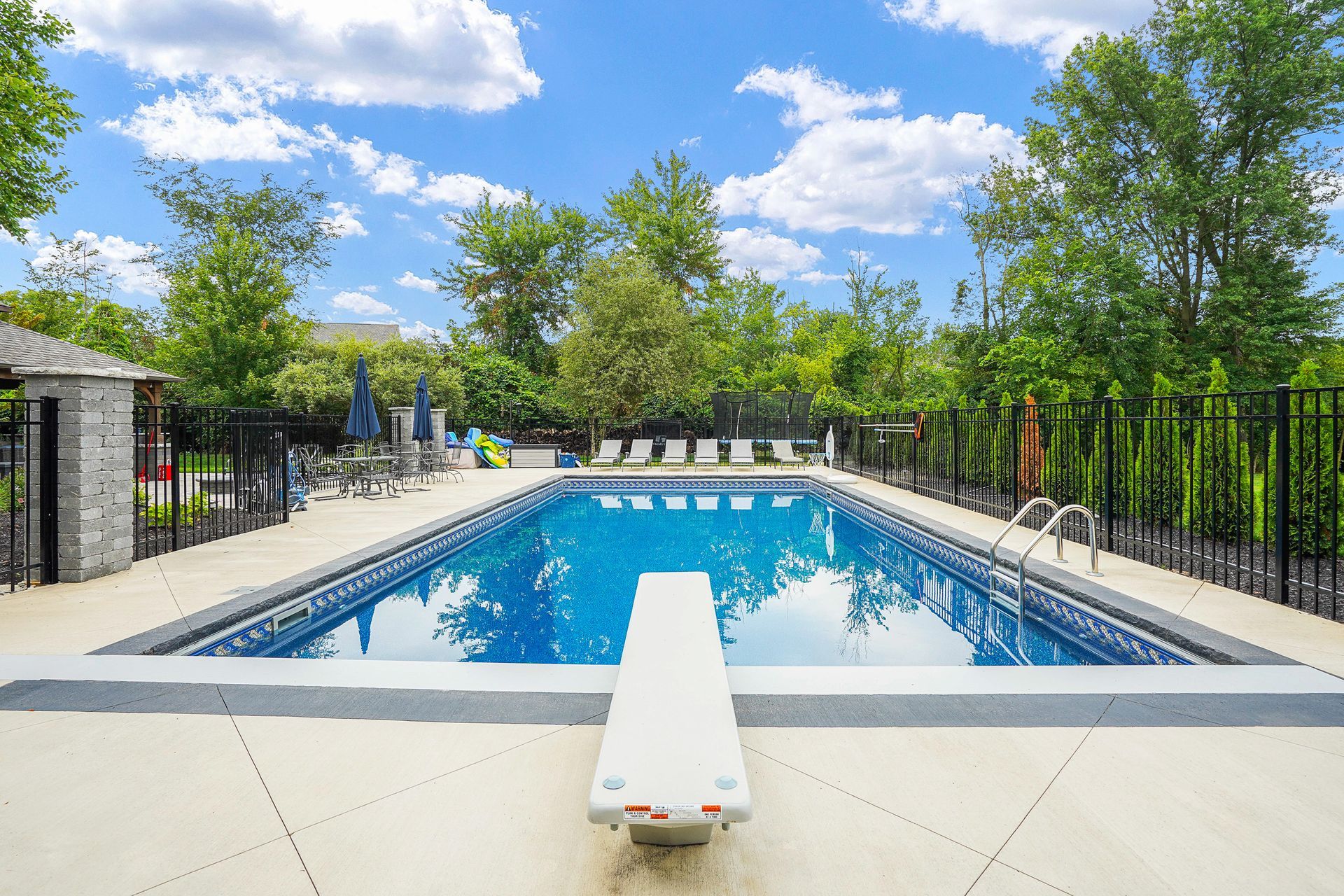 A rectangular outdoor swimming pool with a diving board on a sunny day. Green trees surround the pool area.