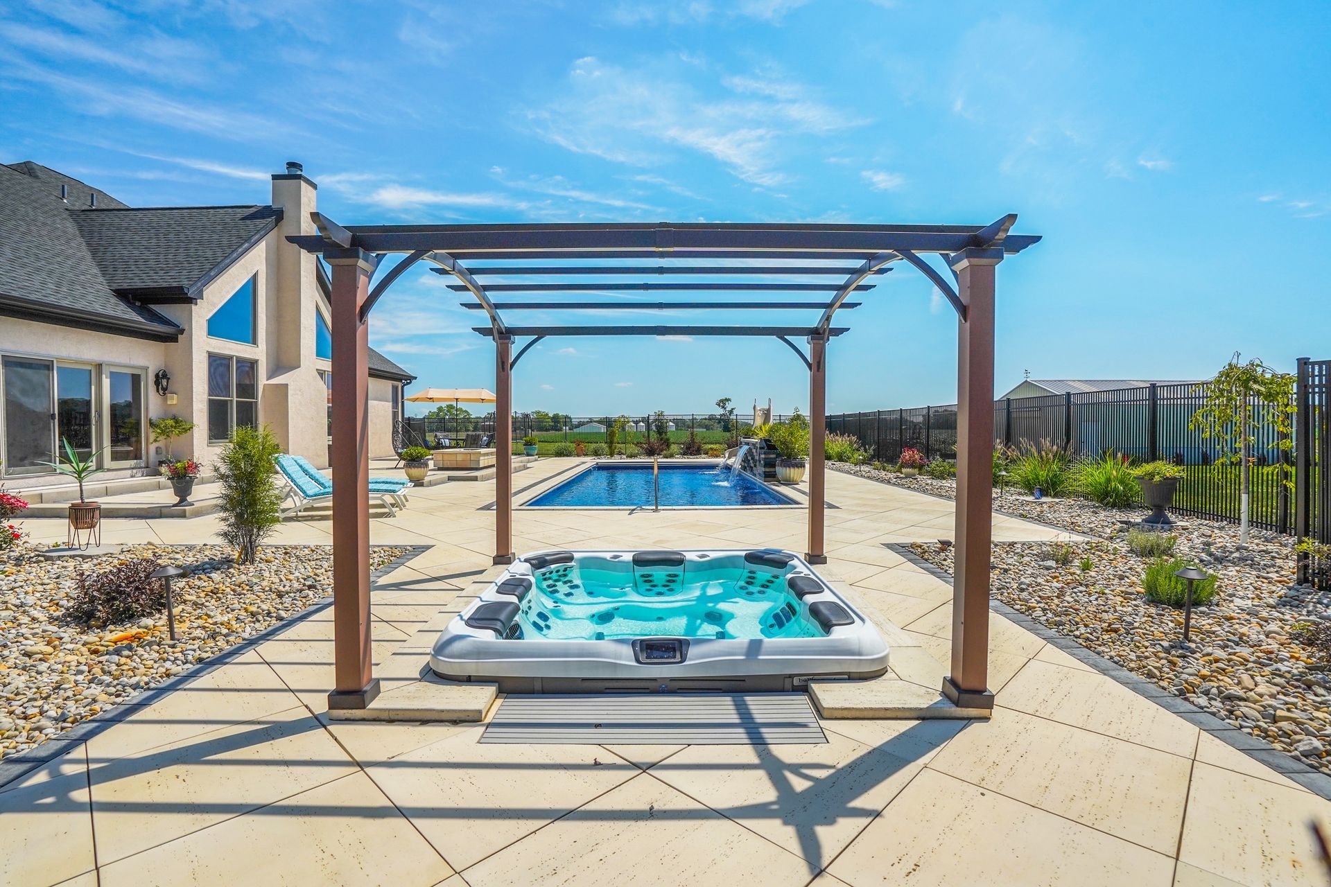A hot tub sits under a pergola on a patio, with a pool and expansive yard in the background.