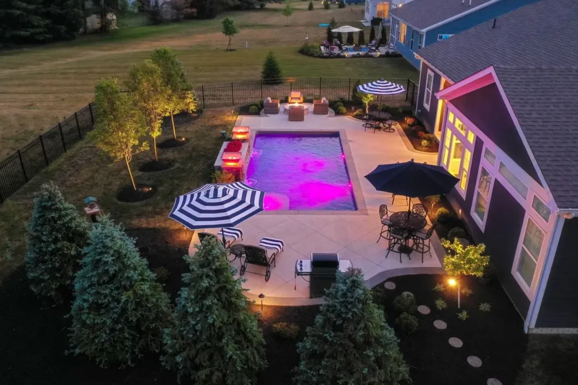 An enchanting nighttime view of a backyard oasis, complete with a sparkling pool and a well-lit outdoor living patio.