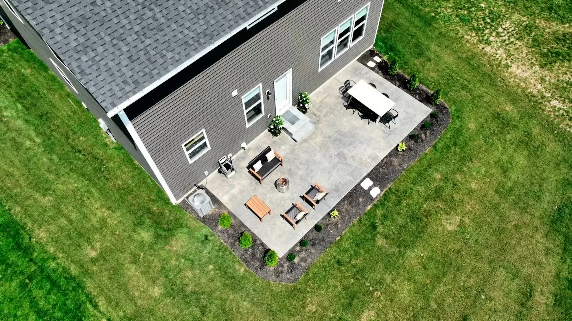 Overhead view of a house with a gray exterior, patio, and green lawn. Patio furniture includes a table and chairs, and a seating area with a fire pit.