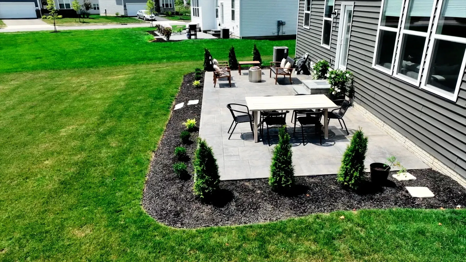 A backyard patio with a dining table and seating area surrounded by landscaping and a green lawn. The house has gray siding.