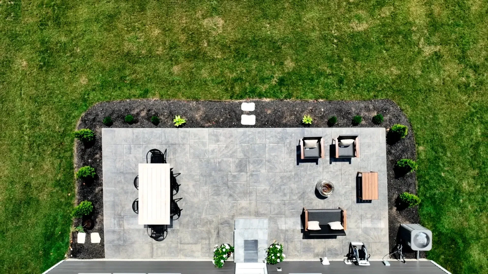 Overhead view of a patio with dark gray pavers, outdoor furniture, a dining table, and a fire pit, surrounded by landscaping and a green lawn.