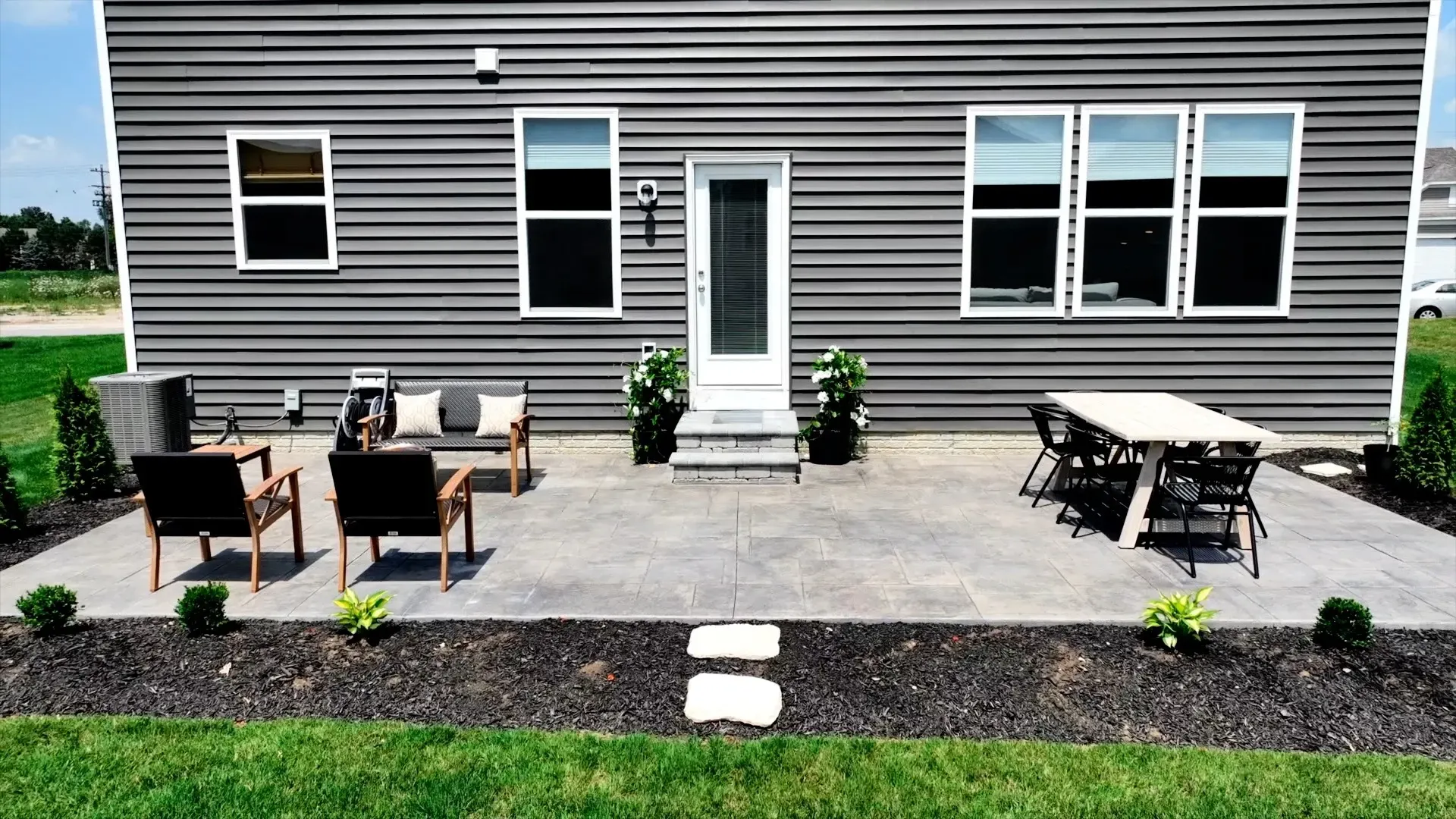 A backyard patio with seating areas, a dining table, and a dark gray house with windows. Green grass surrounds the patio.
