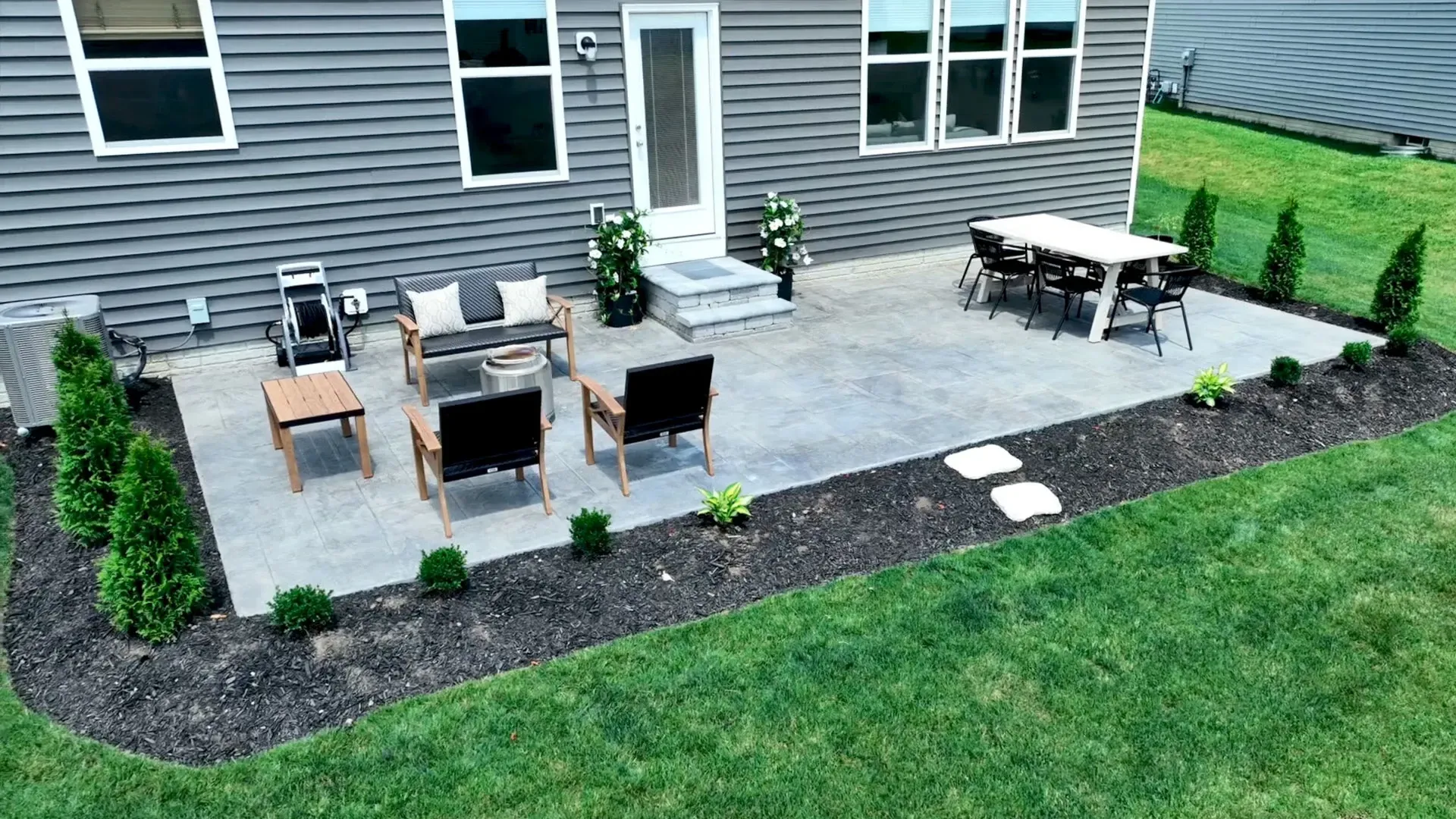 Backyard patio with furniture, including a table, chairs, and a couch, on a gray stone surface. Landscaping with green plants and black mulch surrounds the patio.
