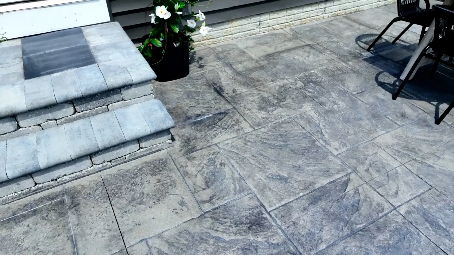 A gray patio with stone pavers, leading to steps and a doorway. A potted plant and shadows of furniture are visible.