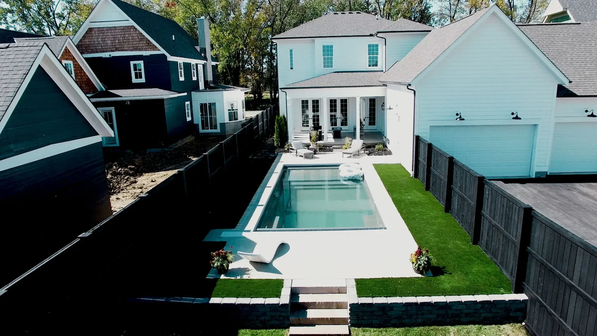 Aerial view of a modern backyard with a rectangular pool, patio, and lush green lawn.  A white house and garage are in the background.