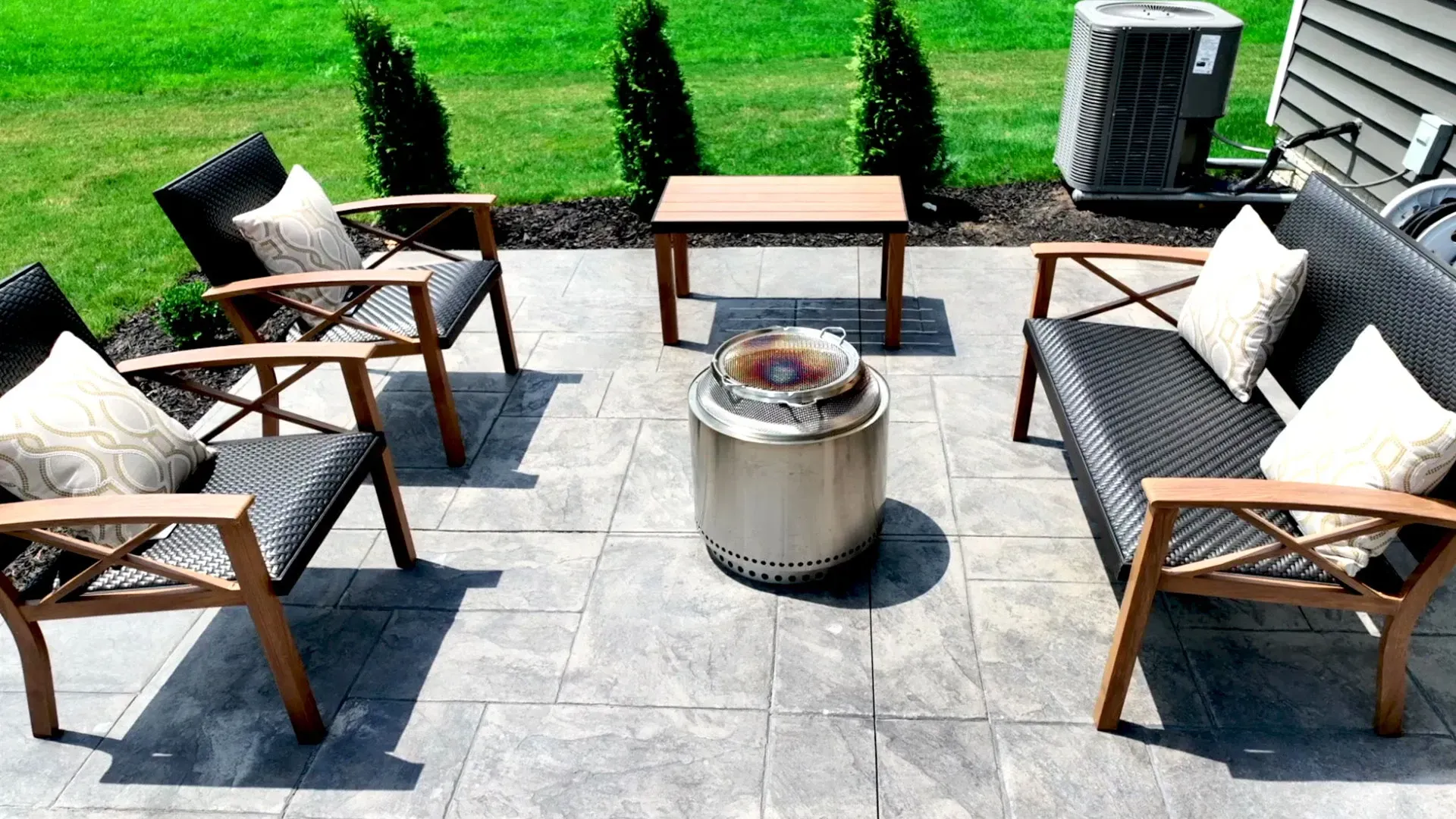 Patio with seating arranged around a stainless steel fire pit.  Includes four chairs, a sofa, a small table, and greenery.