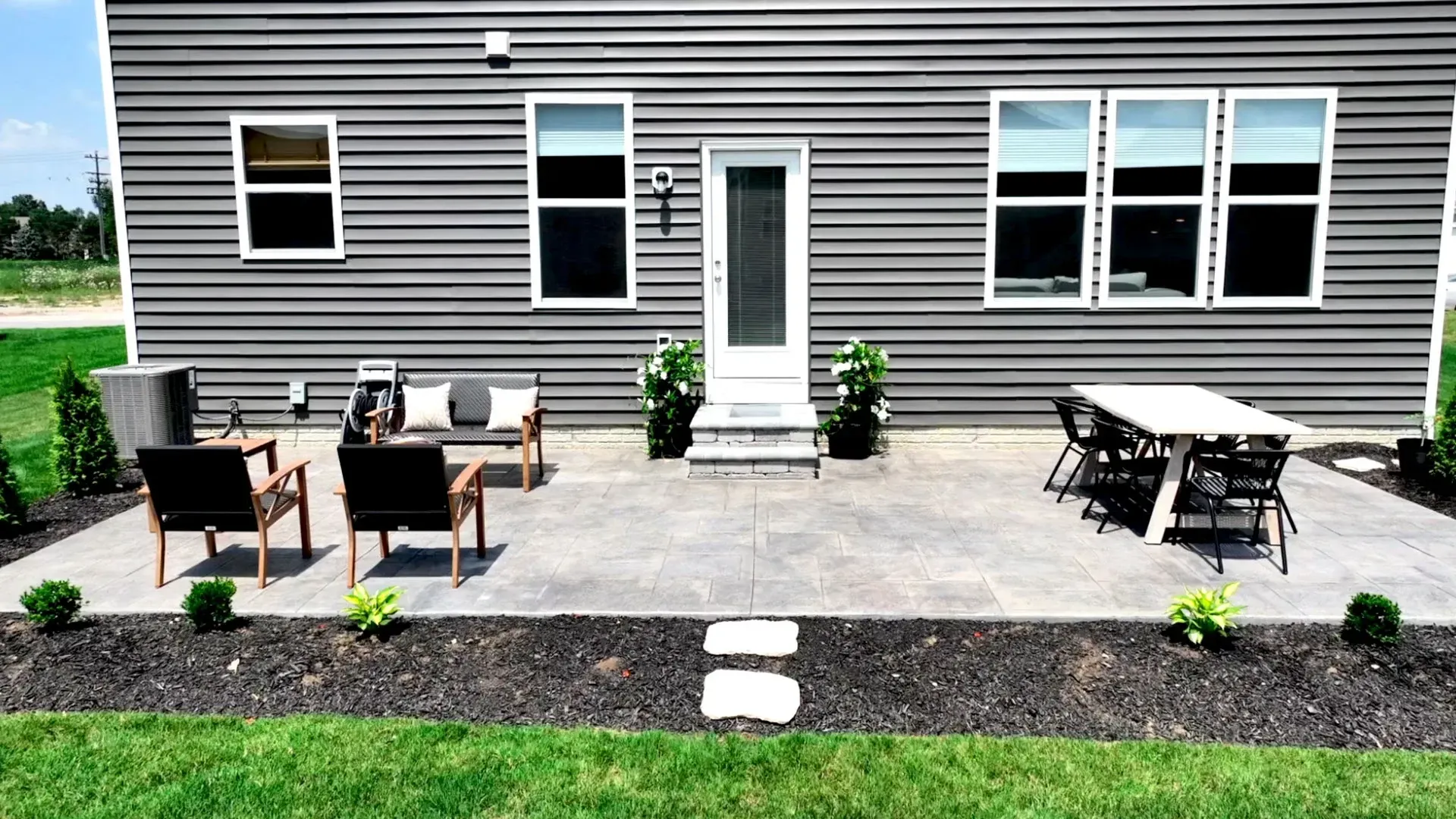 A gray house with a concrete patio furnished with outdoor seating and a dining table, surrounded by landscaping.
