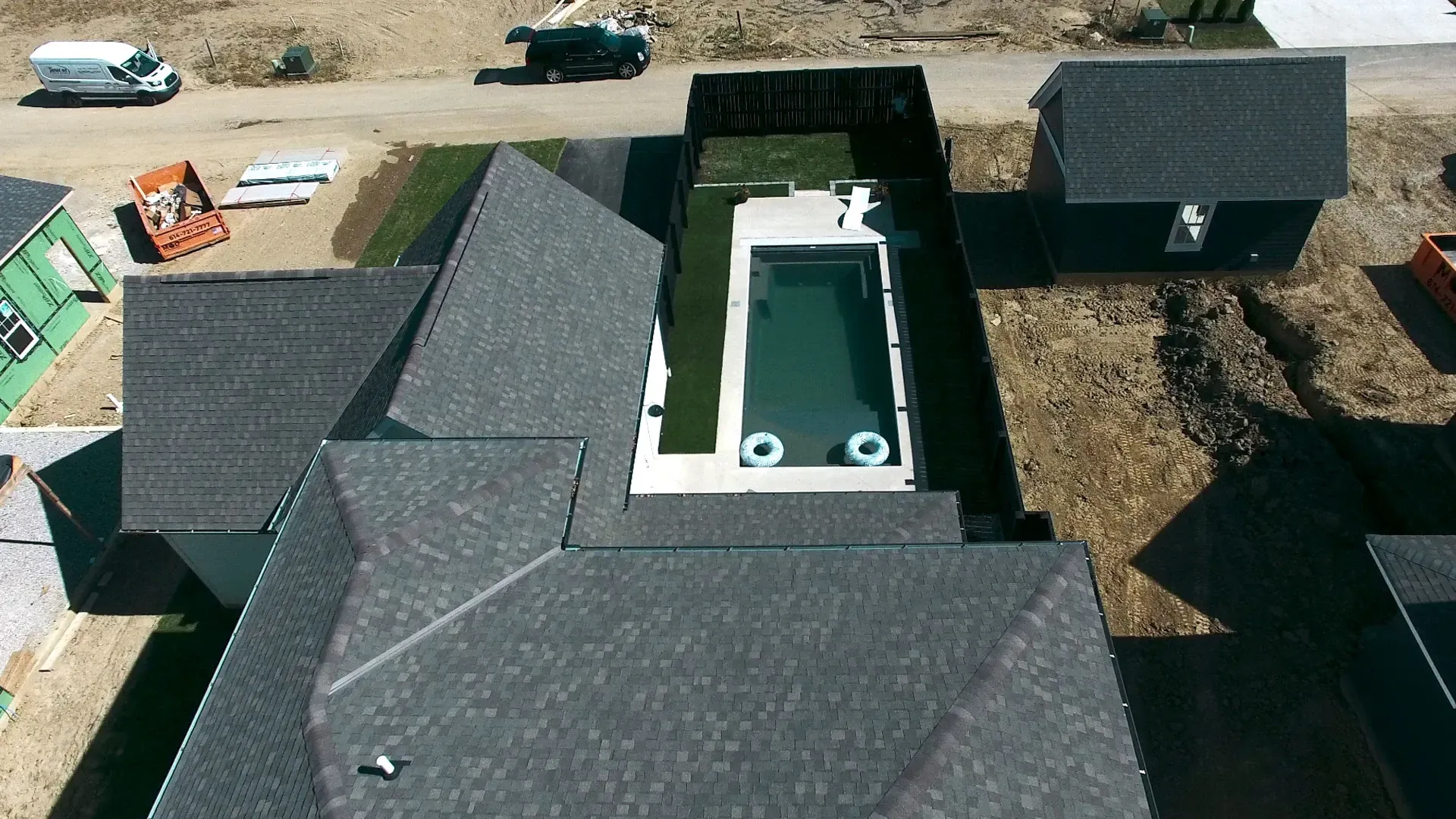 Aerial view of a house with a dark grey roof and a rectangular pool in the backyard. Construction materials are visible in the surrounding area.