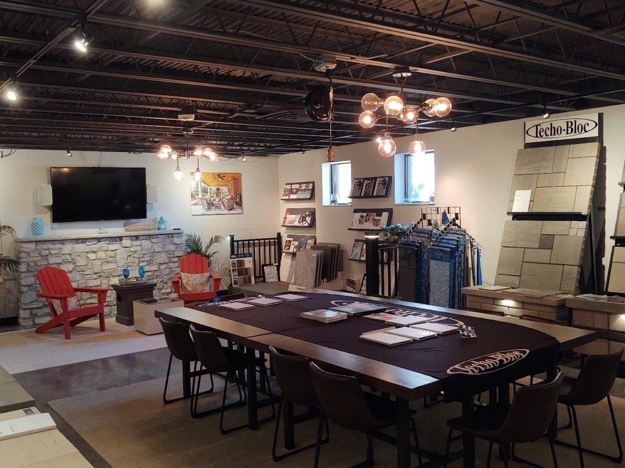 A showroom with stone displays, a large table, chairs, and a seating area with a fireplace, TV, and red chairs.