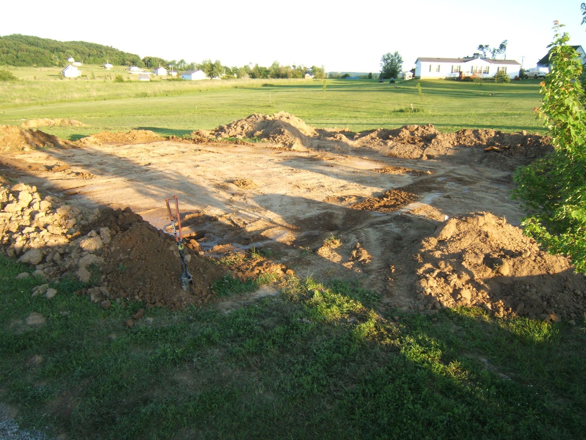 A cleared area of earth with piles of dirt around the perimeter, likely a construction site in a grassy field.