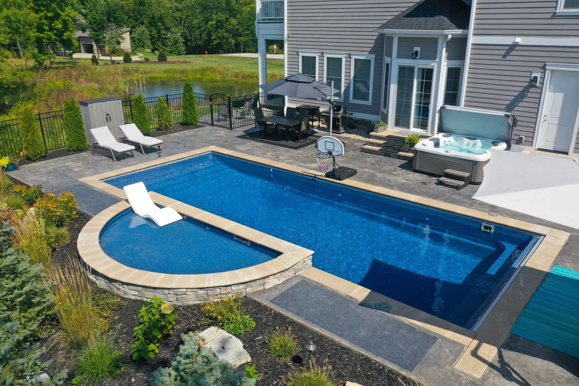 Aerial view of a backyard with a blue tiled pool, hot tub, lounge chairs, and a house with gray siding.