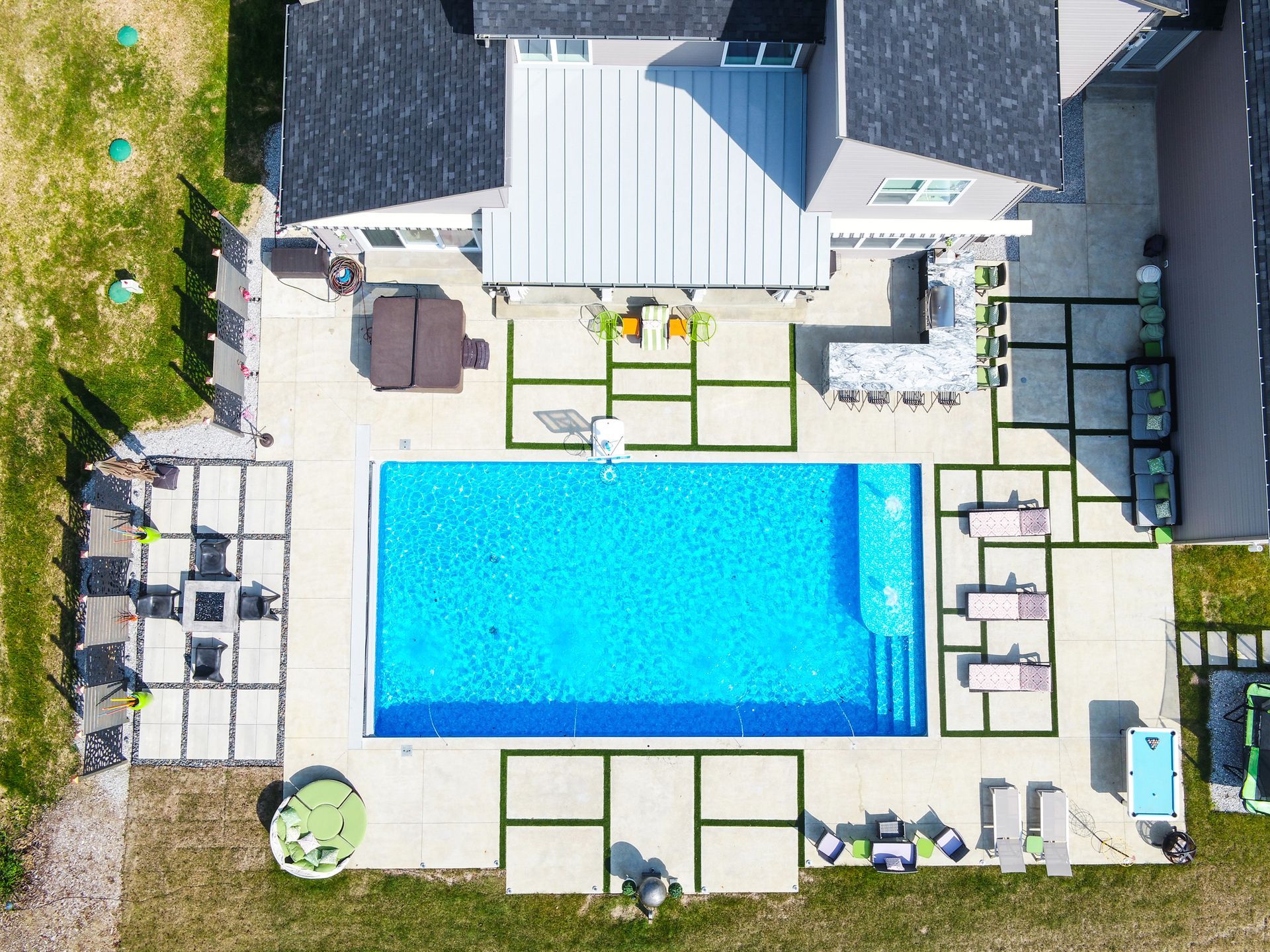 Aerial view of a backyard with a rectangular pool, patio, and lawn. A house, lounge chairs, and bar seating are visible.