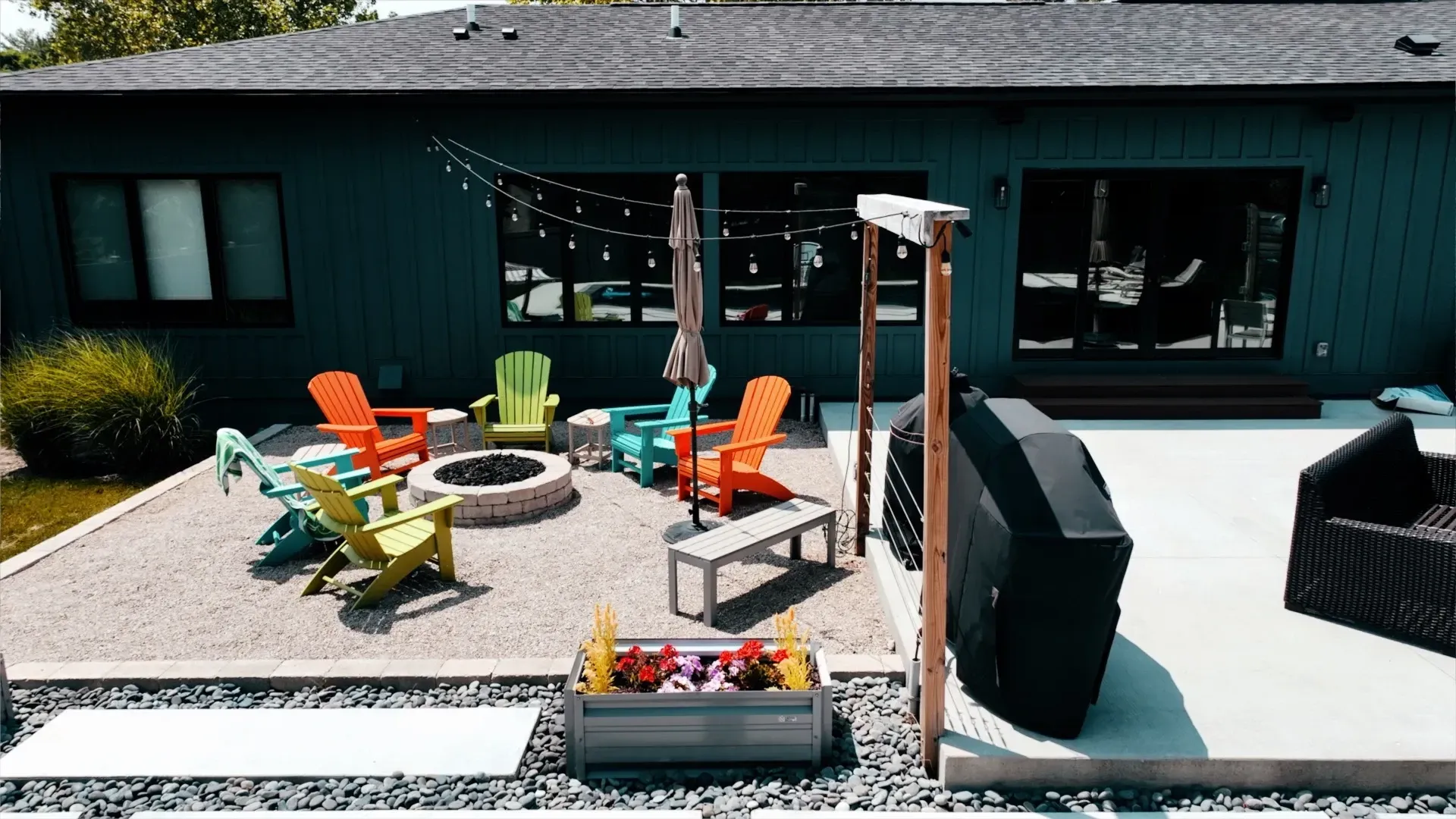 Backyard patio with colorful Adirondack chairs around a fire pit. String lights, a bench, and a grill are also visible.