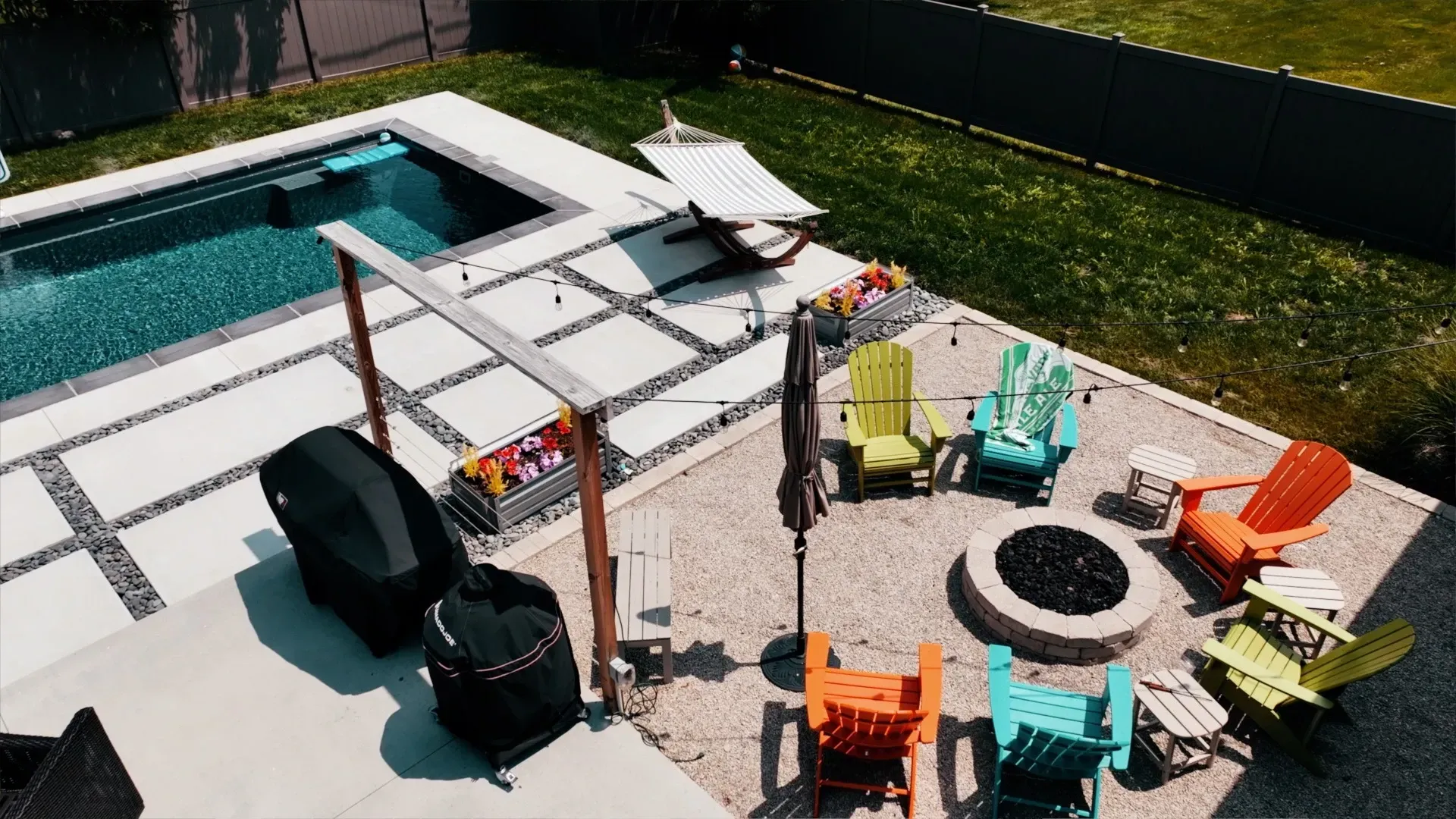 Backyard scene with a pool, fire pit surrounded by colorful chairs, and grilling area.