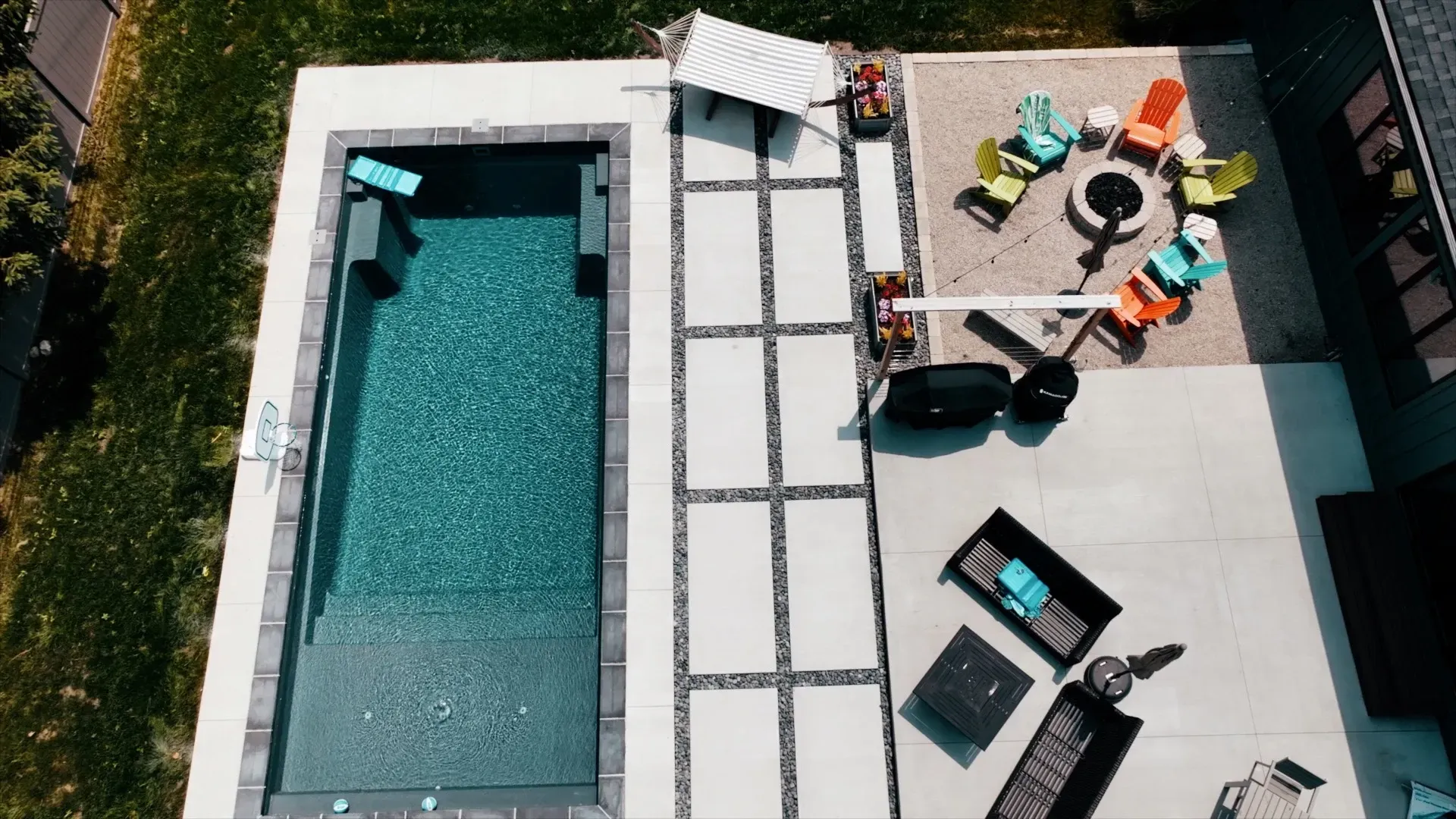 Aerial view of a backyard with a pool, seating area with a fire pit, and colorful Adirondack chairs on a gravel patio.