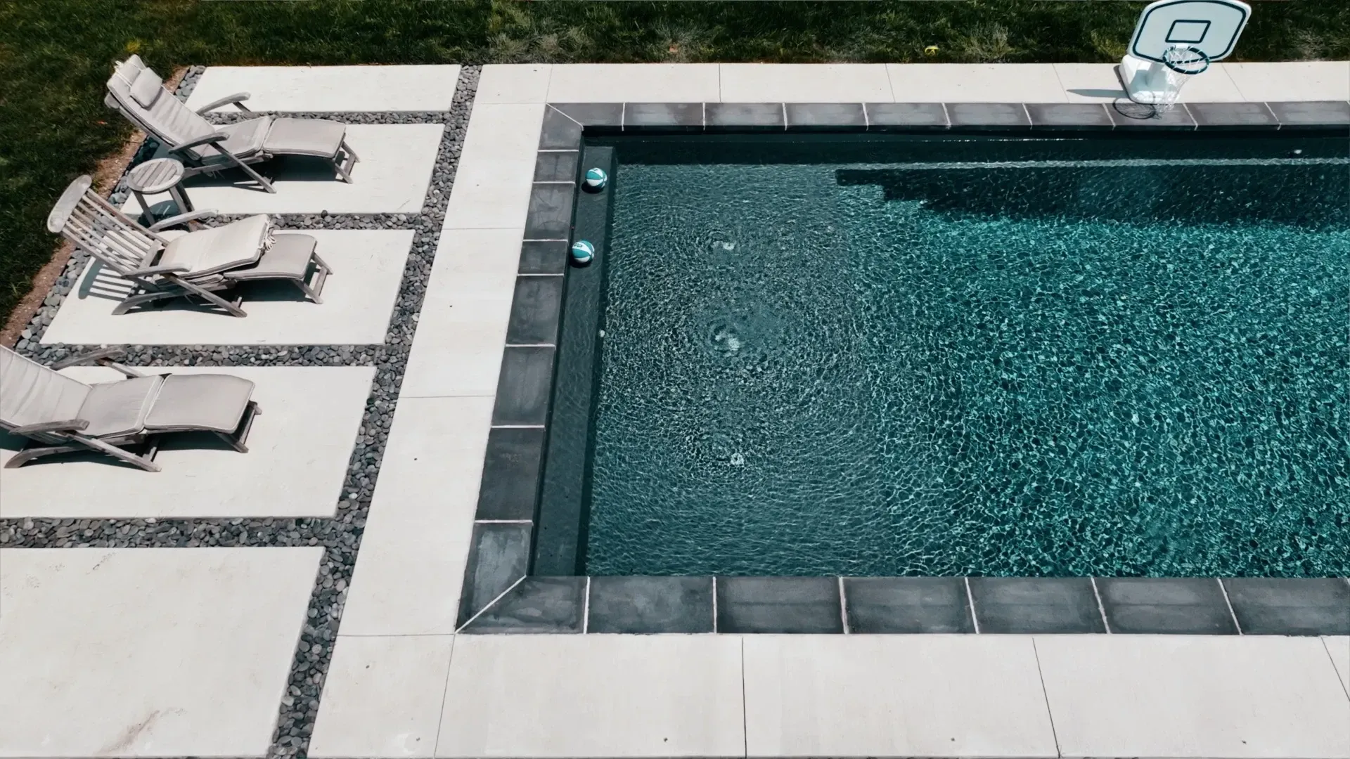 Overhead view of a turquoise pool with a dark tile border, surrounded by a concrete patio and lounge chairs.