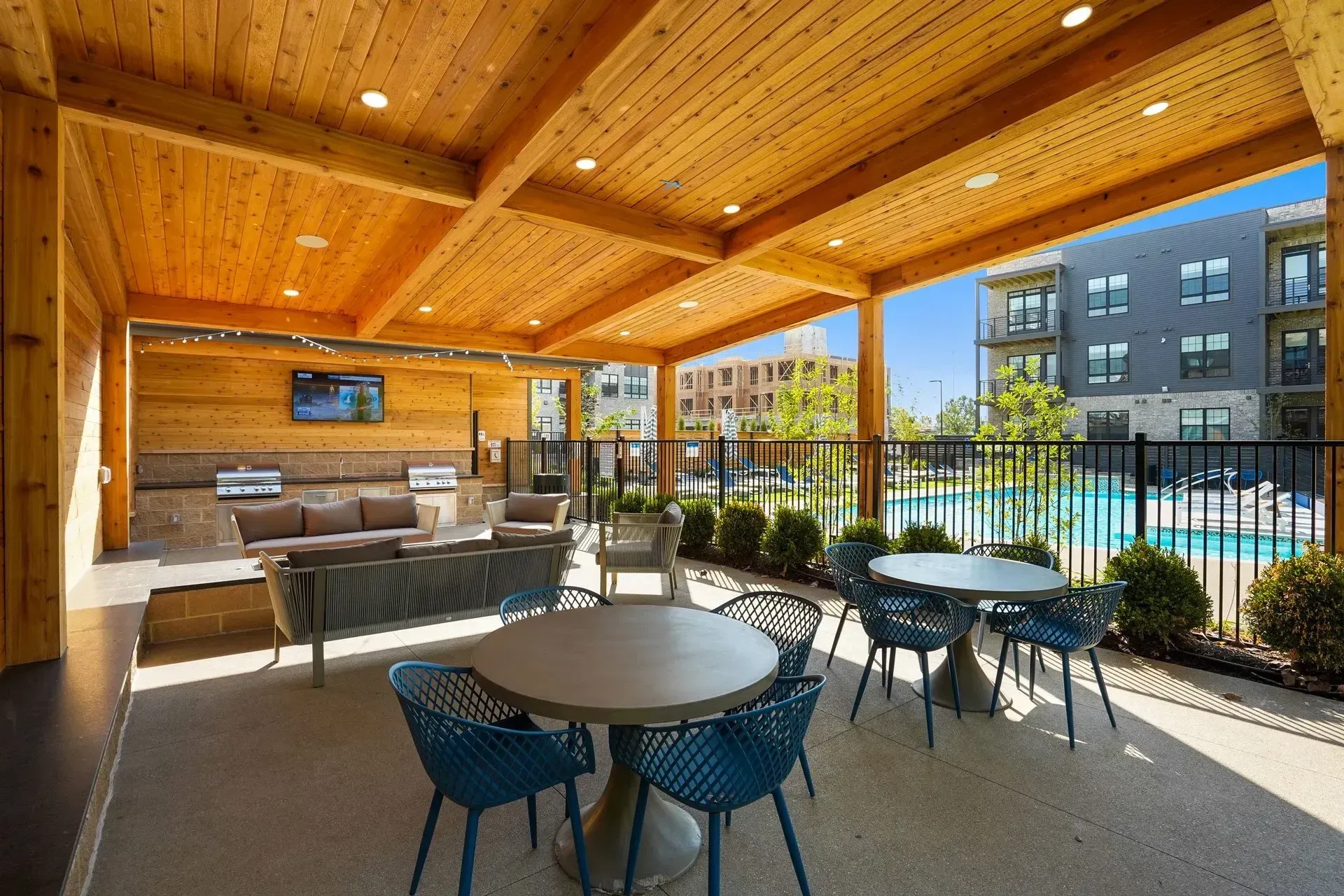 Outdoor patio with seating, a grill, and a TV, adjacent to a pool and apartment building. Features wood ceilings and blue chairs.
