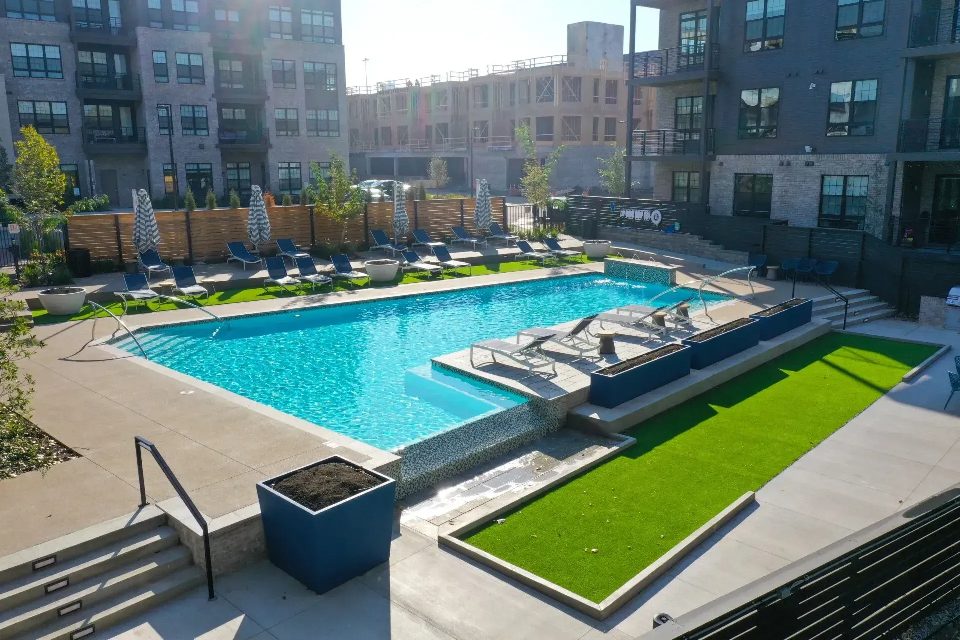 Apartment complex pool area with turquoise water, lounge chairs, and green turf. Buildings surround the pool under a sunny sky.