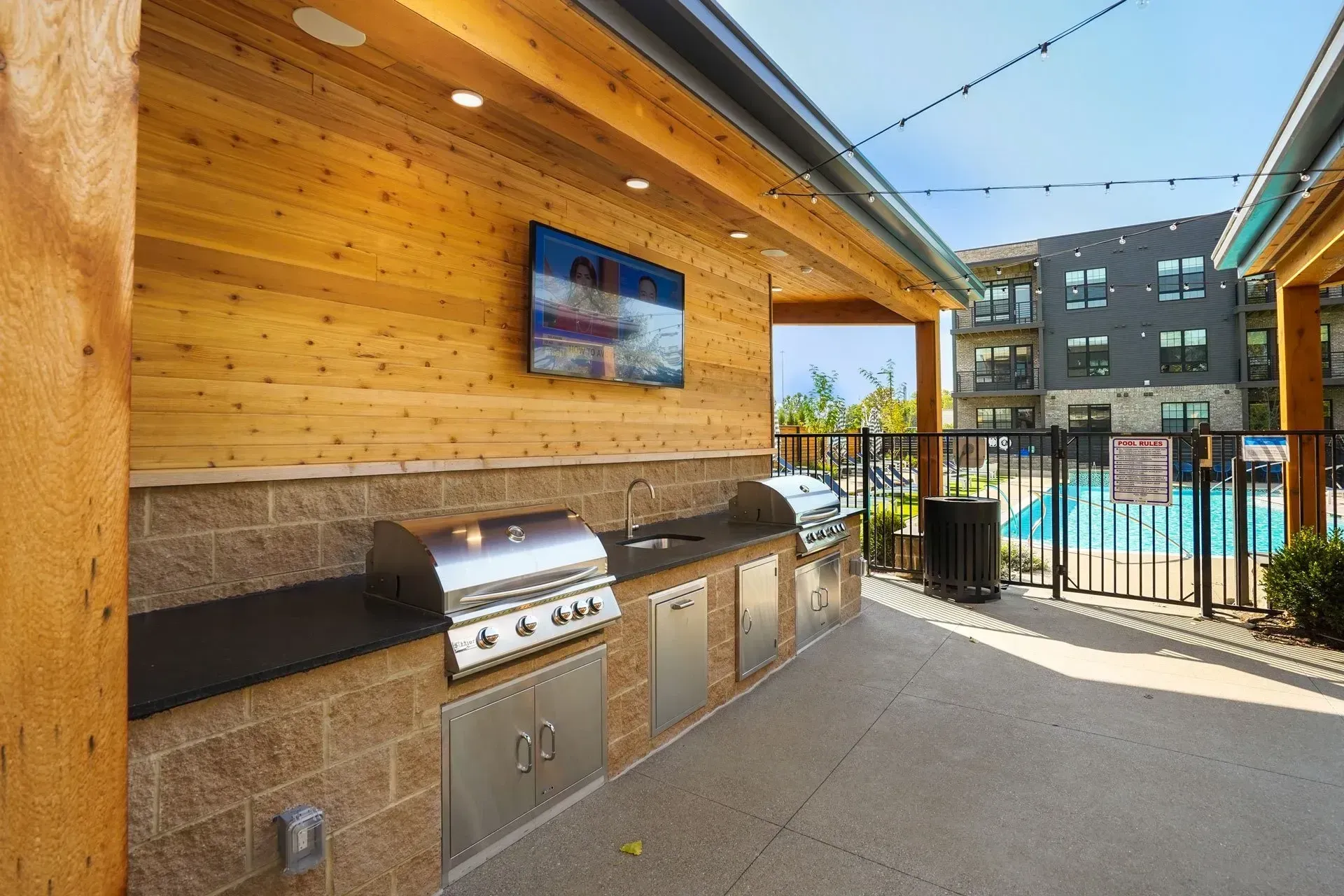 Outdoor kitchen area with grills, sink, TV, and seating near a pool.