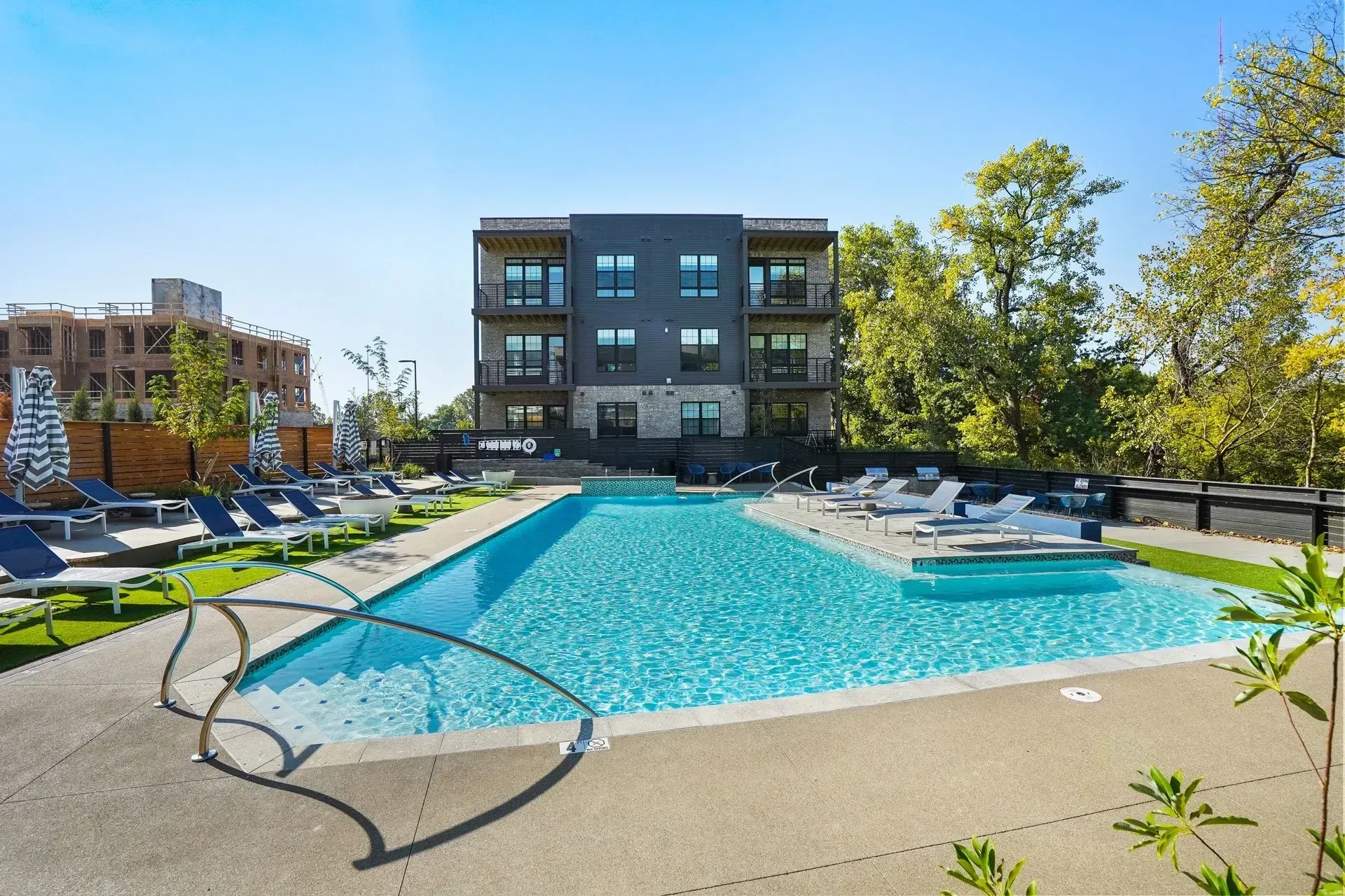 An apartment complex with a pool and lounge chairs on a sunny day. A three-story building sits in the background.