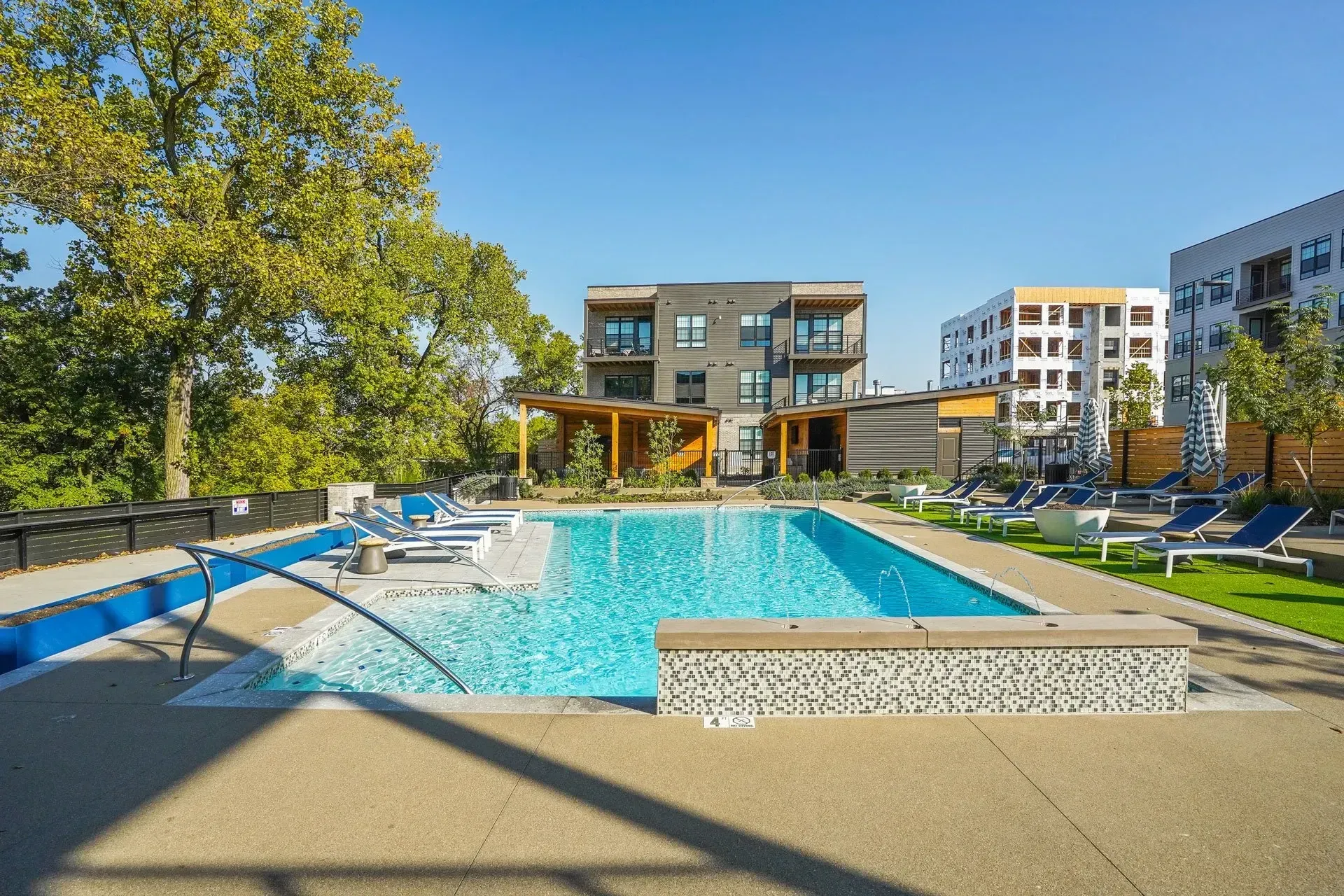 A rectangular pool in a modern apartment complex, with lounge chairs, and buildings in the background under a sunny sky.