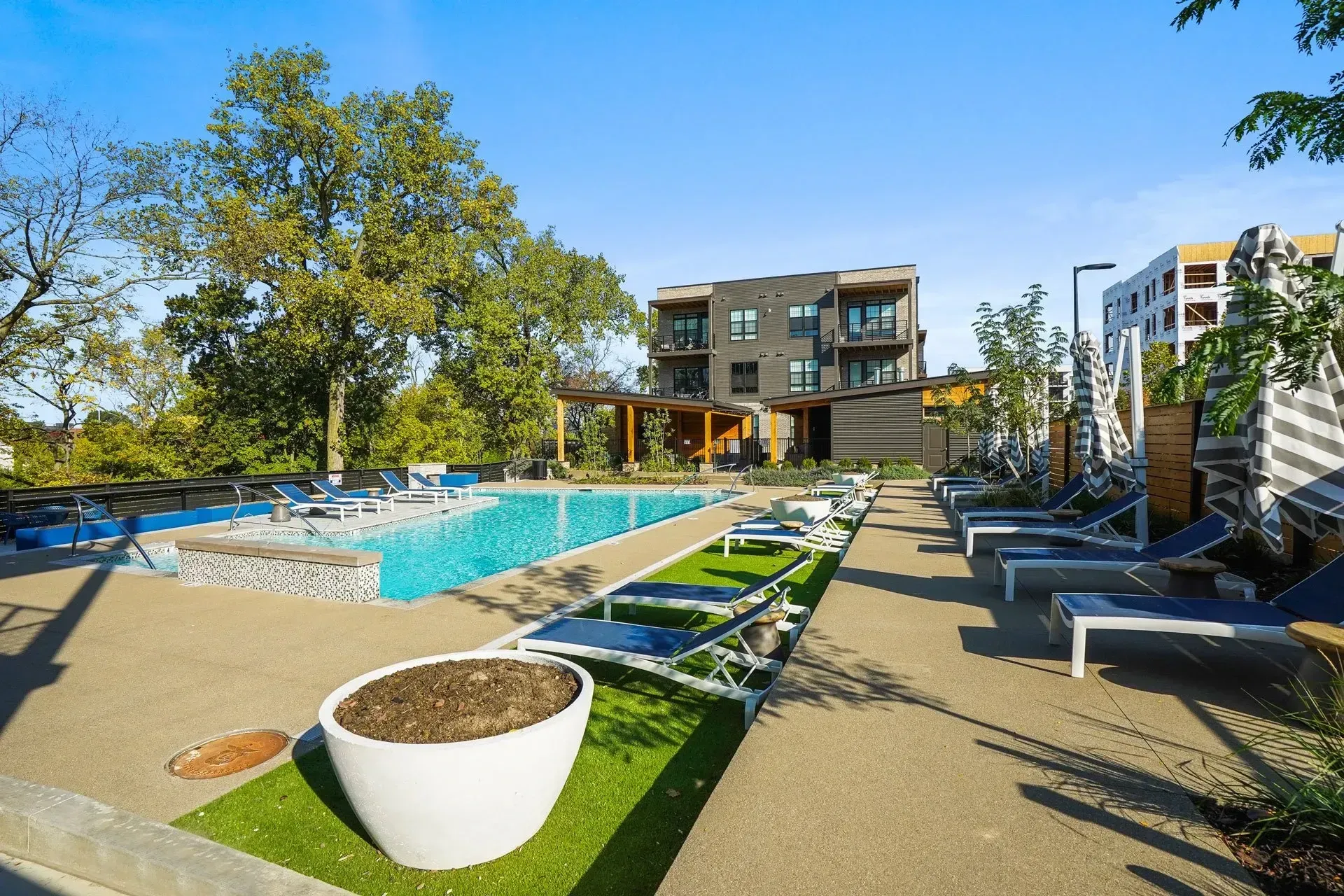 Pool area with lounge chairs, a modern building, and trees on a sunny day. A large white planter sits in the foreground.