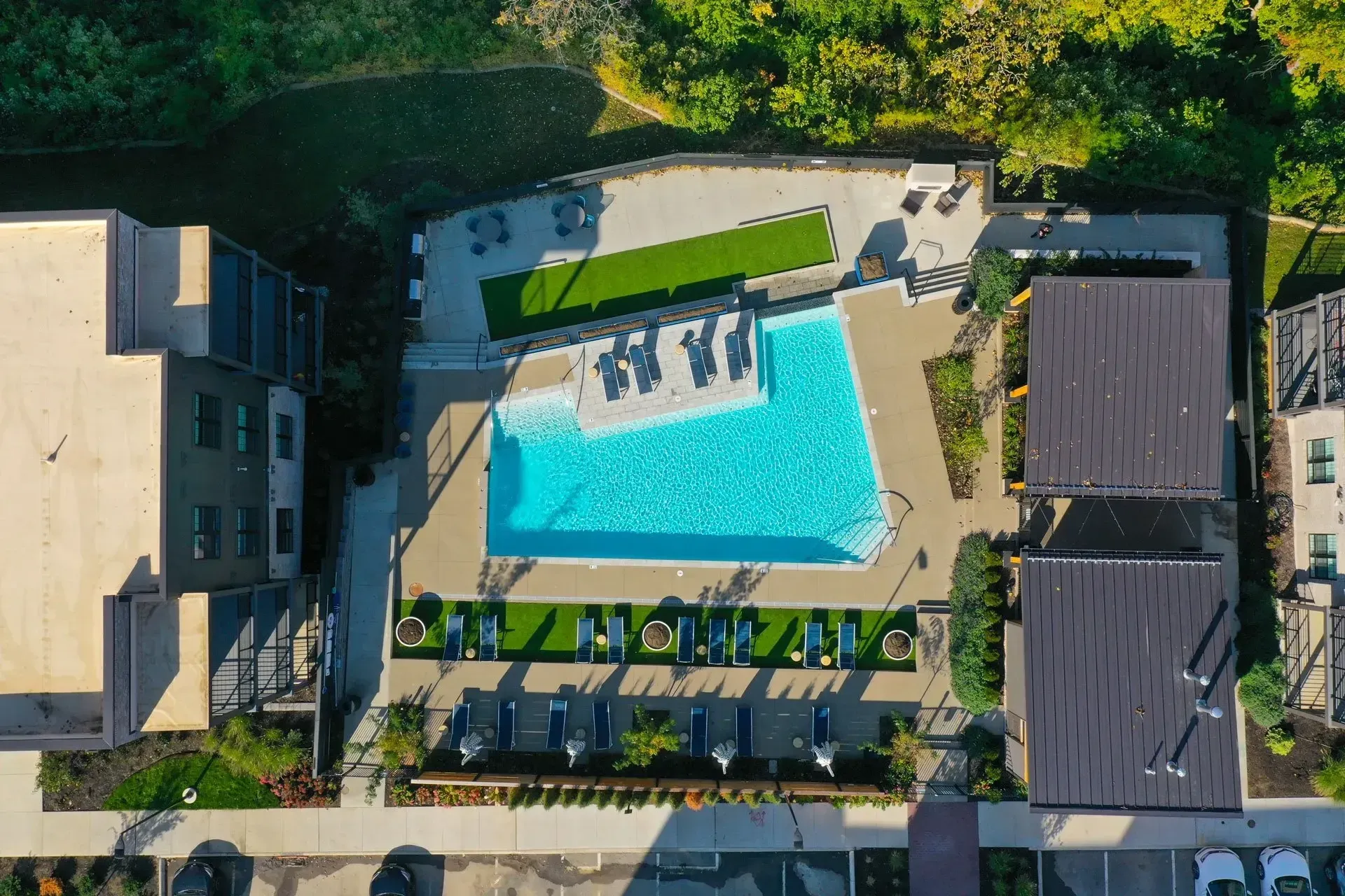 Overhead view of a turquoise swimming pool surrounded by patio seating, artificial grass, and modern buildings.