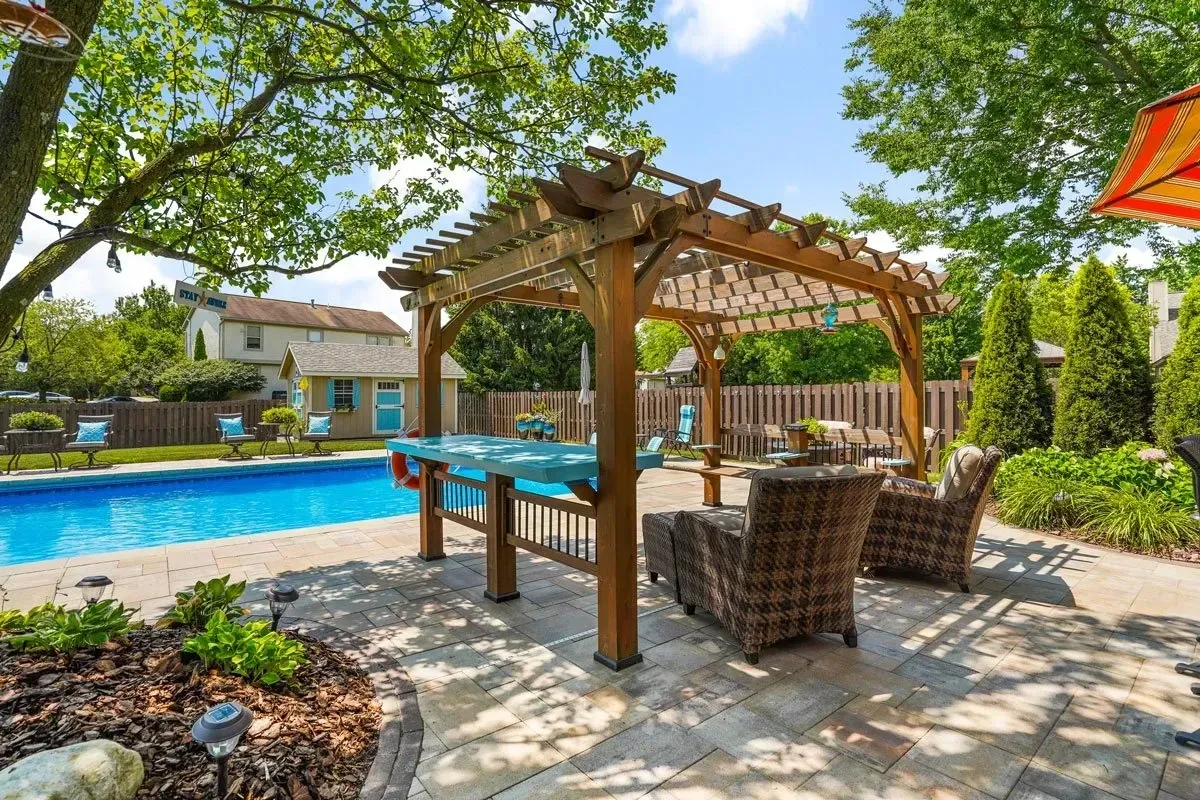 Outdoor patio with a pergola over a blue table and seating, adjacent to a pool. Lush greenery surrounds the patio.