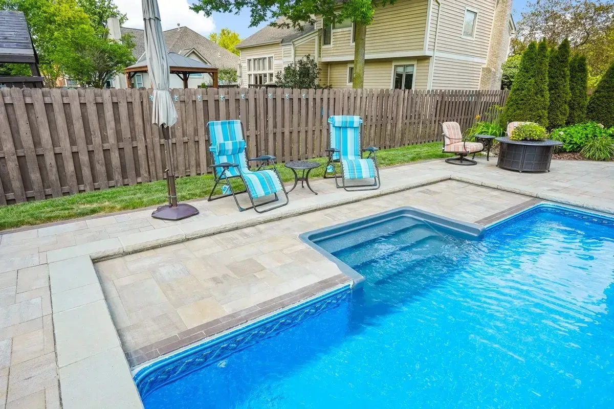 Backyard pool with blue water, surrounded by a paved patio area, fence, and lounge chairs.