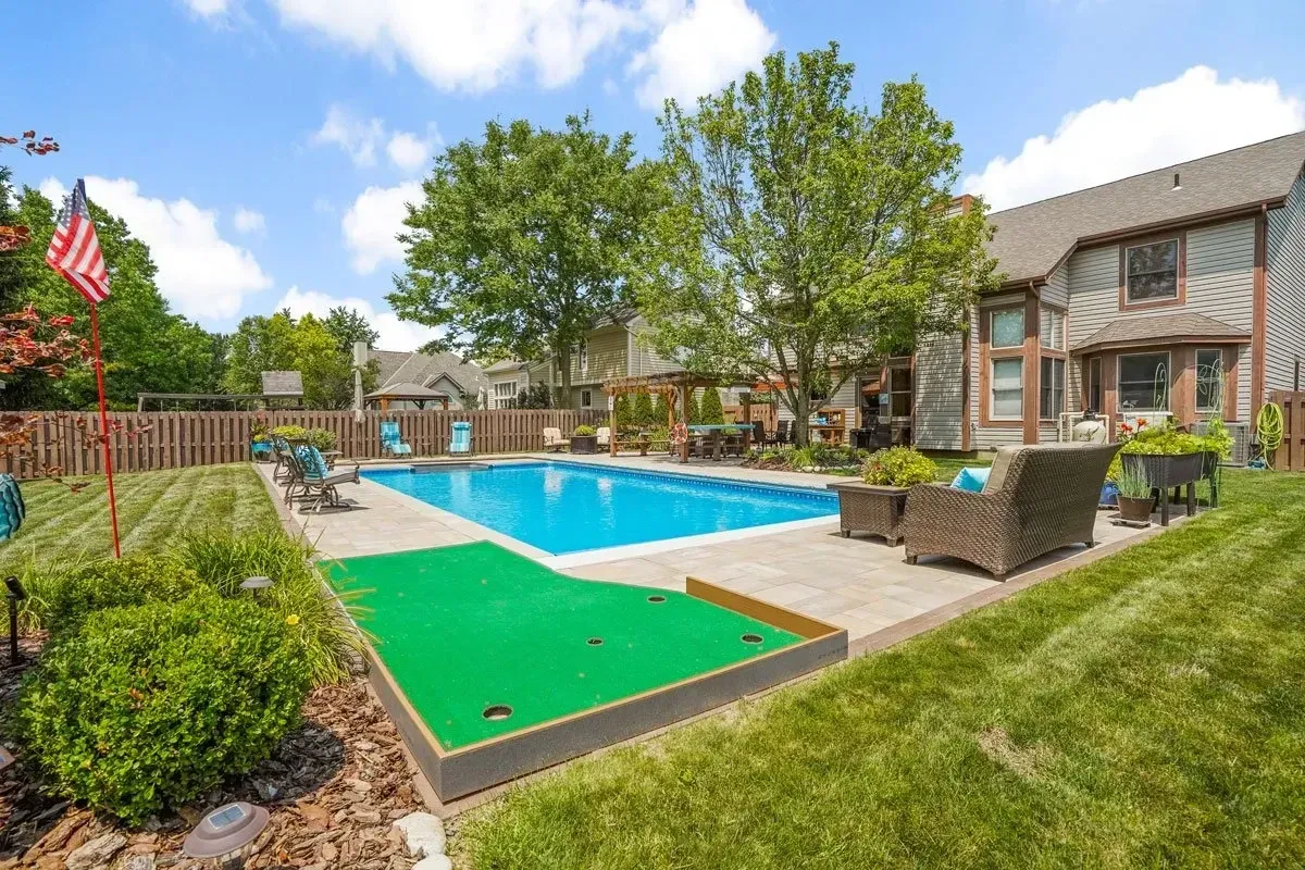 Backyard with swimming pool, putting green, patio furniture, and a two-story house with trees and blue sky. An American flag waves in the background.