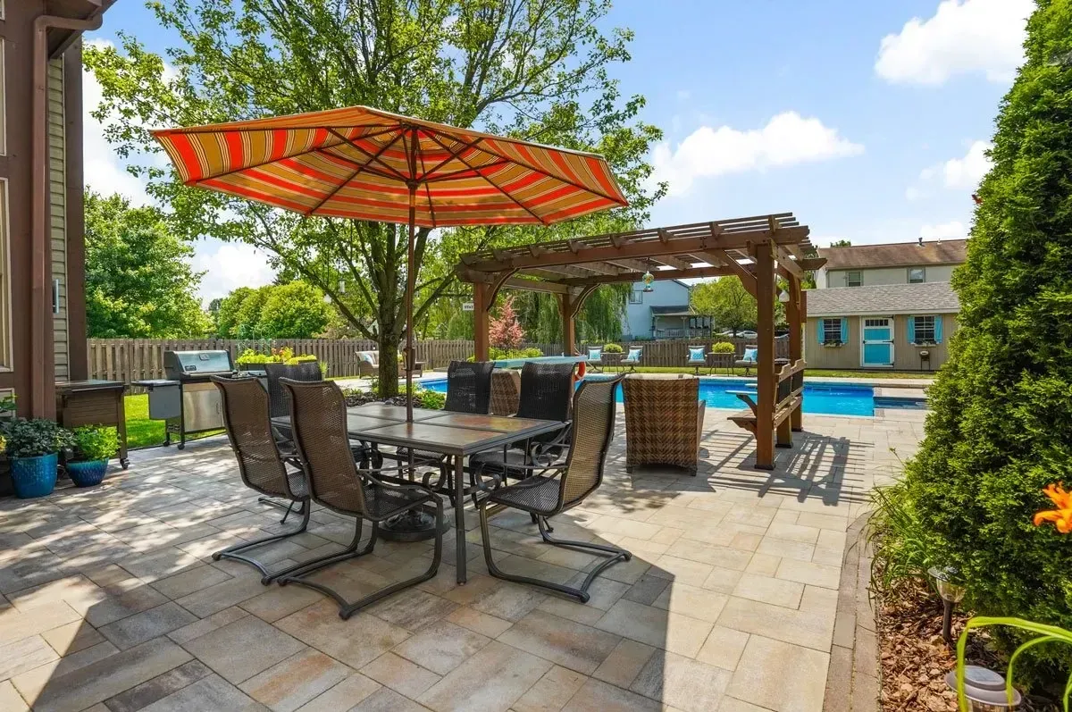 Patio with outdoor dining table, chairs, grill, and umbrella, beside a swimming pool and pergola on a sunny day.