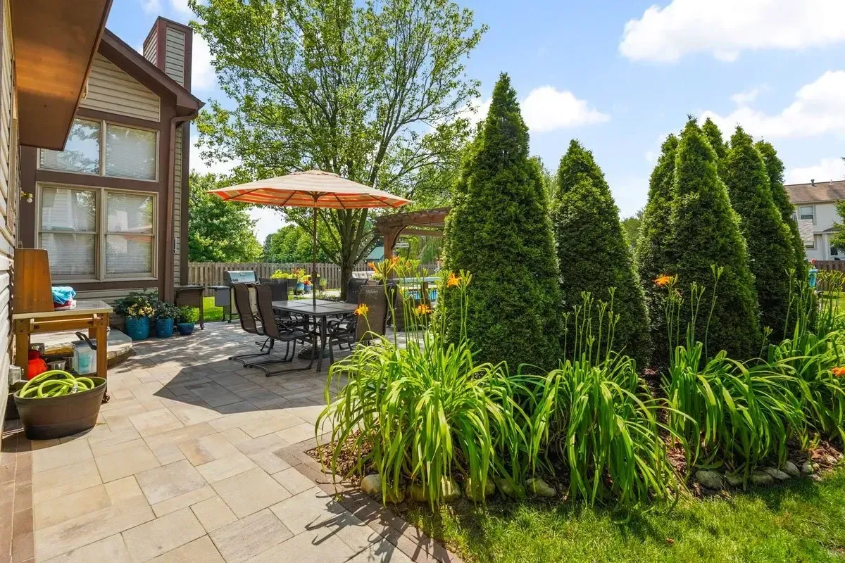 Patio with stone pavers, a dining set under an umbrella, and evergreen trees. Green grass and blue sky in the background.
