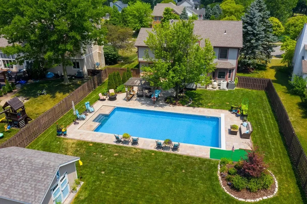 Aerial view of a backyard with a rectangular pool, seating area, and a two-story house surrounded by green grass and a wooden fence.
