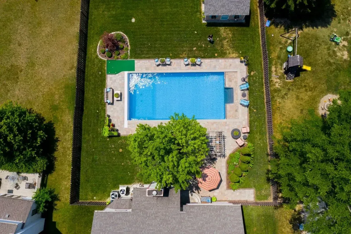 Aerial view of a backyard with a rectangular pool, patio, green lawn, and a large tree in the center.