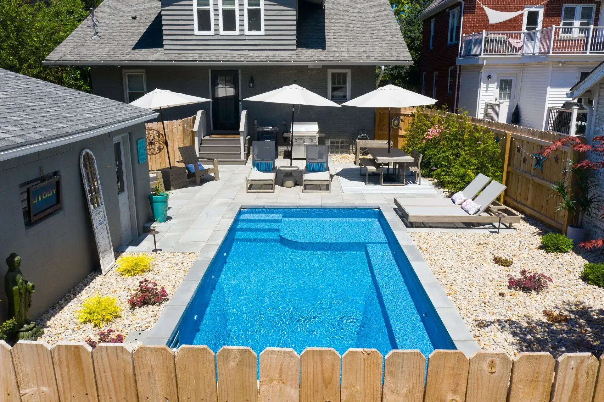 Backyard with a blue rectangular pool, patio furniture, and a light gray house. A shed is to the left, and a wooden fence in the foreground.