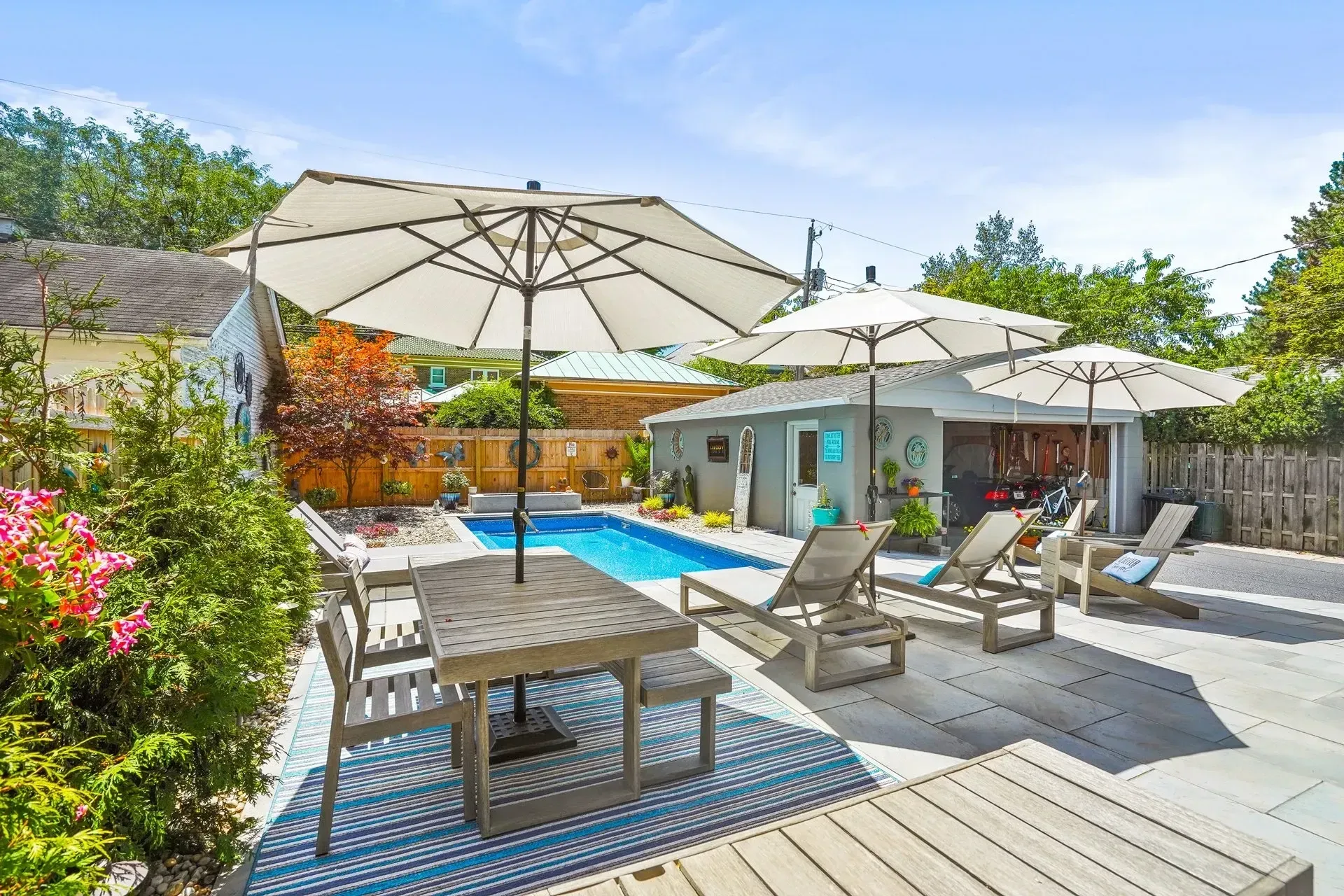 Backyard with a pool, patio, and lounge chairs under umbrellas. A wooden table with chairs is also present.