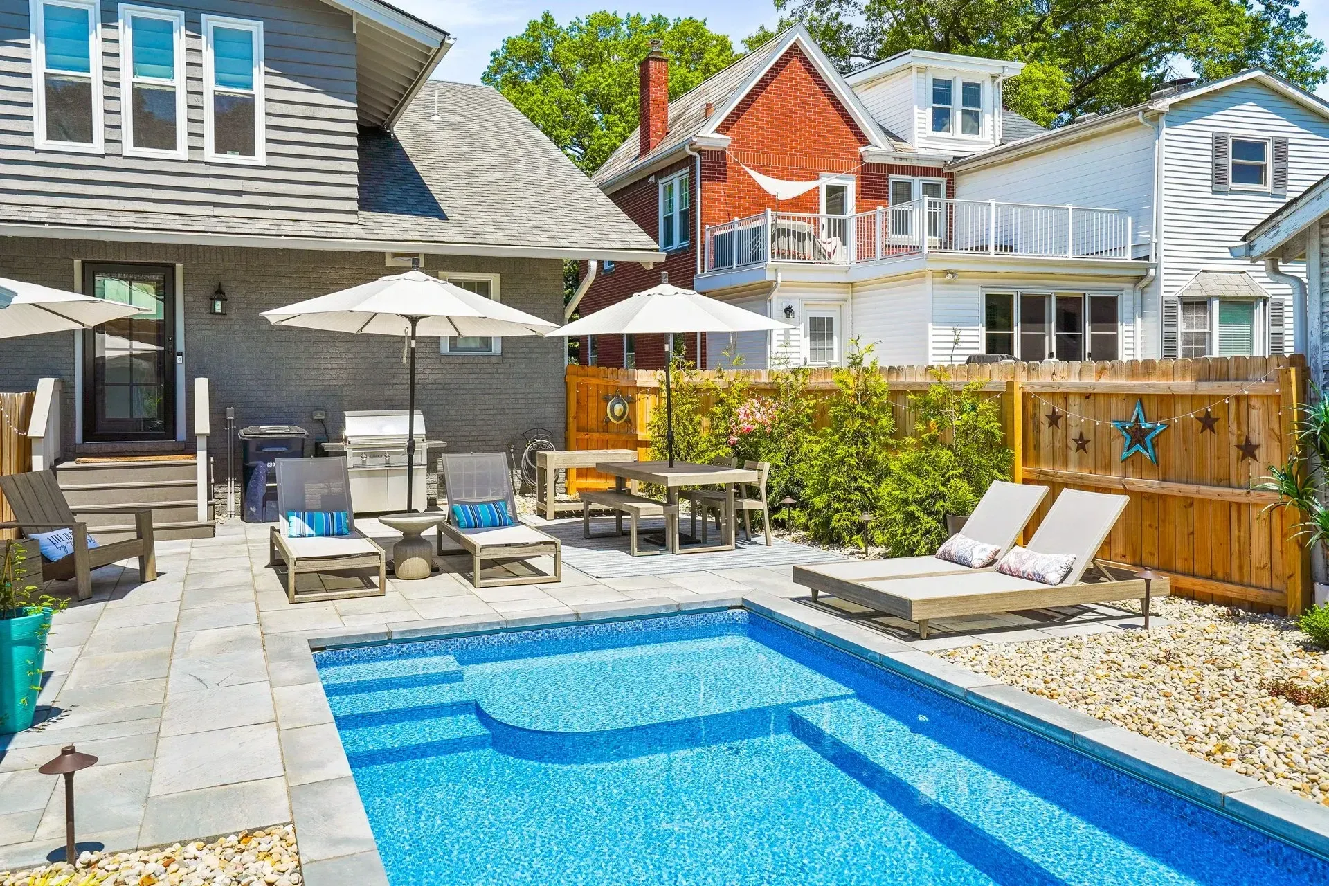 Backyard with a pool, lounge chairs, and a dining area. Houses with gray and red exteriors surround the space on a sunny day.