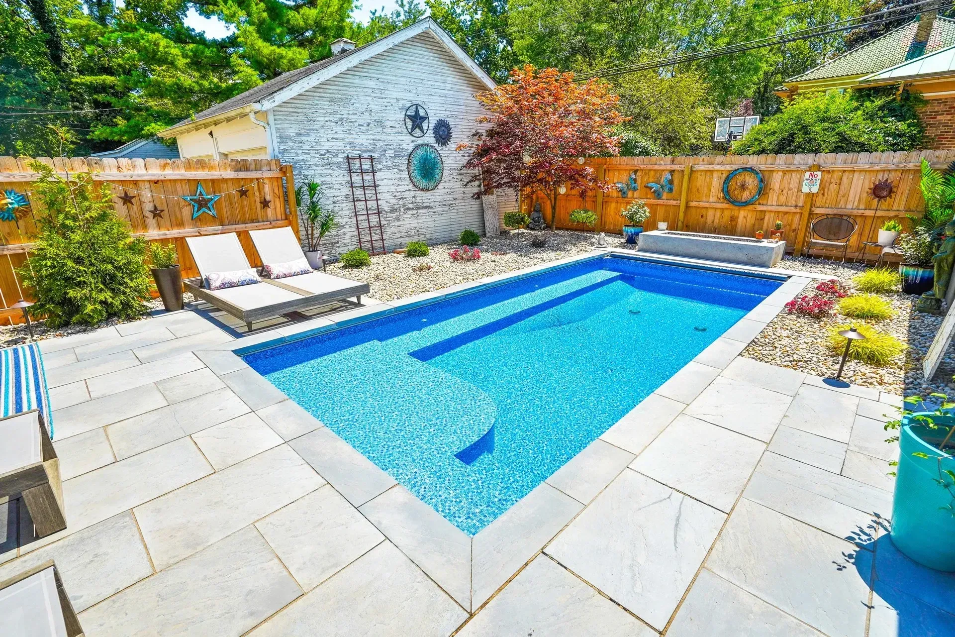 A backyard pool surrounded by stone pavers and a wooden fence. Two lounge chairs sit near the pool, with landscaping and a small building in the background.