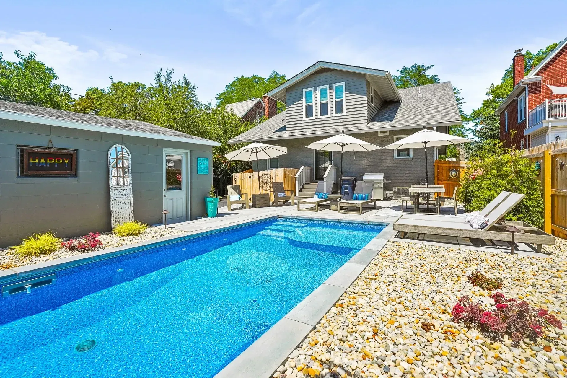 Backyard with a rectangular pool, lounge chairs, umbrella-covered seating, and a gray house.