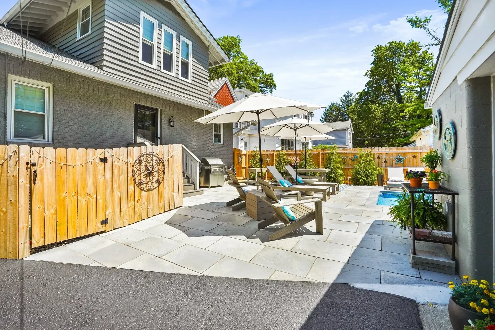 A backyard patio with lounge chairs and umbrellas next to a house, with a wooden fence on the left.