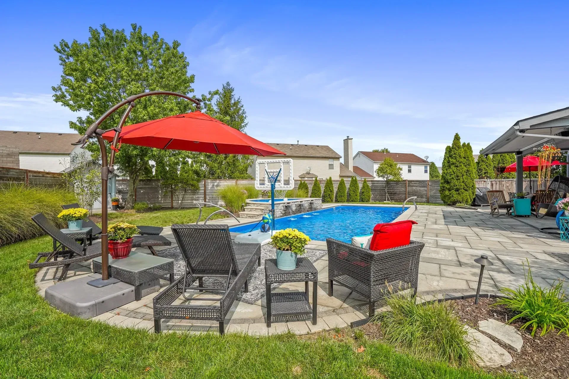 Backyard with a pool, patio, and seating under a red umbrella. Green grass and trees surround the area.