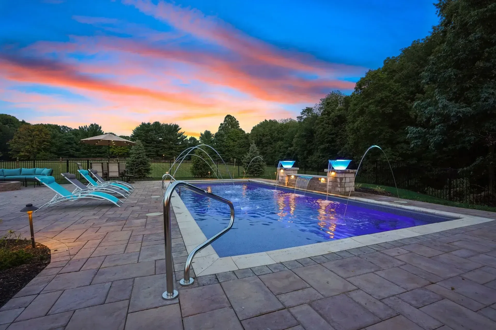 A rectangular pool with water features, surrounded by a stone patio with lounge chairs, under a colorful sunset sky.