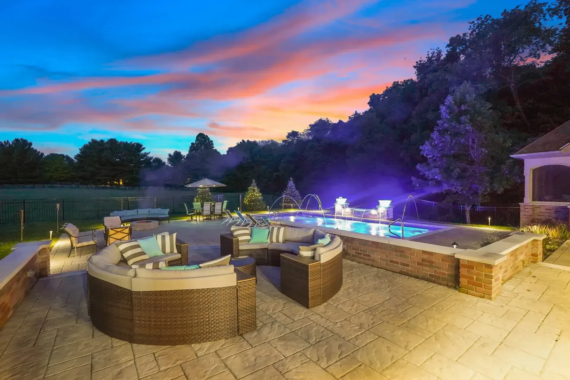 Outdoor patio with pool and seating at dusk. Sunset colors in the sky, pool lit with blue lights, and a wicker seating area.