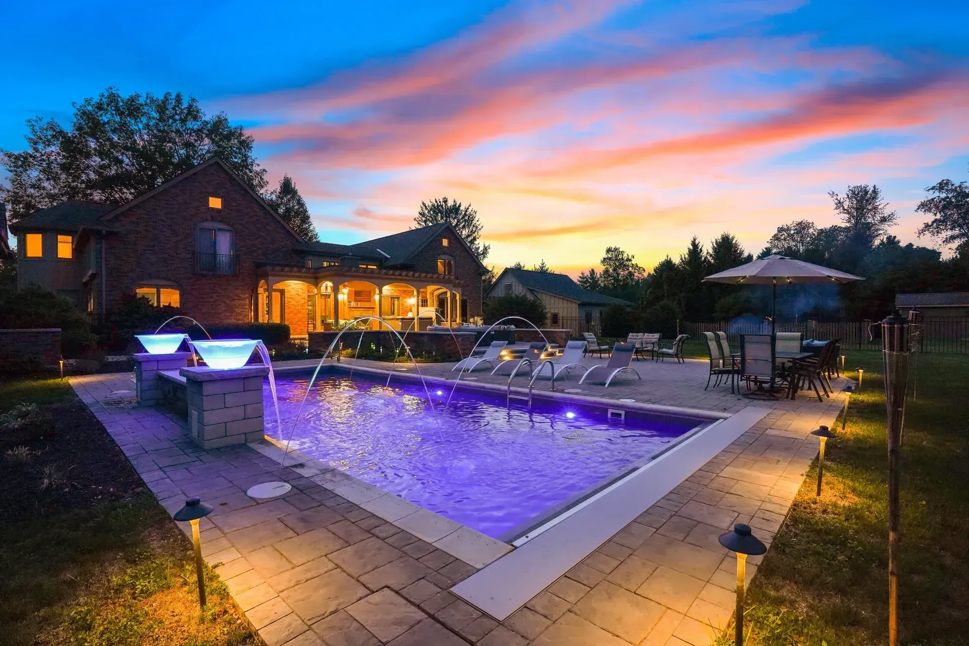 Poolside twilight scene with a brick house, rectangular pool illuminated purple, fountains, and landscape lighting against a colorful sunset.